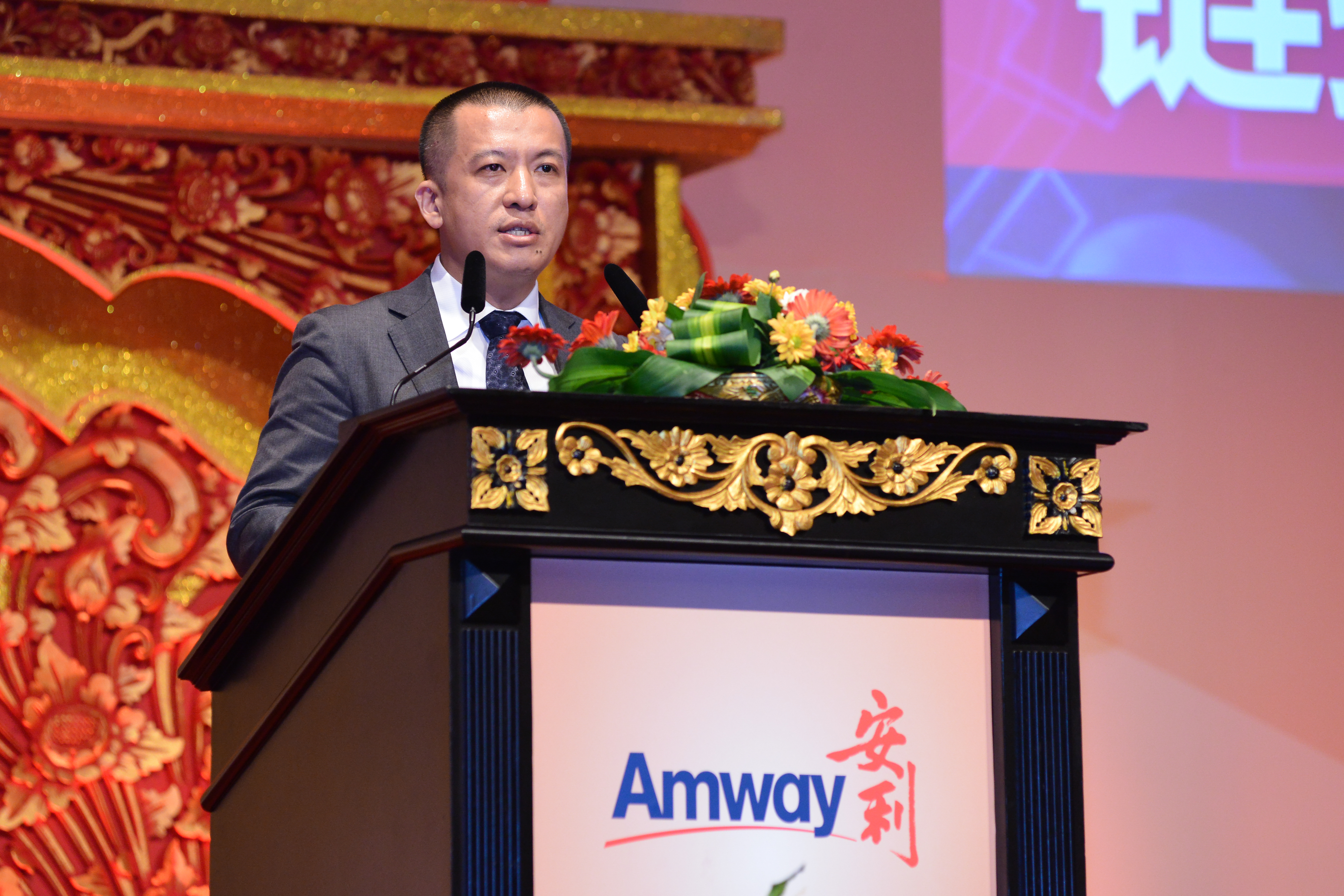 本次安利品牌活动现场，身着正装的高管立于带有“Amway 安利”标识的精美雕花讲台后致辞，讲台摆放着色彩鲜亮的花艺装饰，背景搭配中式雕花装饰与大屏，现场氛围庄重正式，充分展现安利品牌活动的专业风貌。