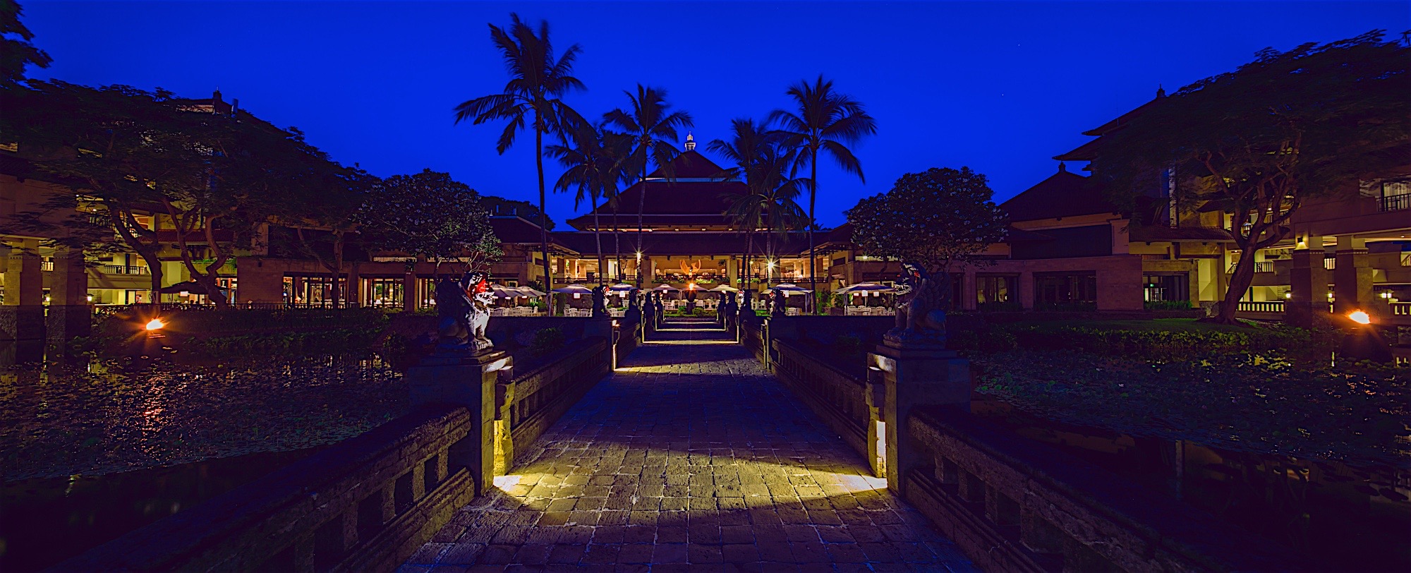 夜幕垂临，场地天空呈浓郁湛蓝色，石板步道连通会场主体，两侧荷塘映着暖光，南洋风格建筑搭配高大椰树，整体氛围静谧雅致、格调高端，尽显安利海外研讨会的高品质办会标准，为参会伙伴营造舒适尊贵的活动环境。