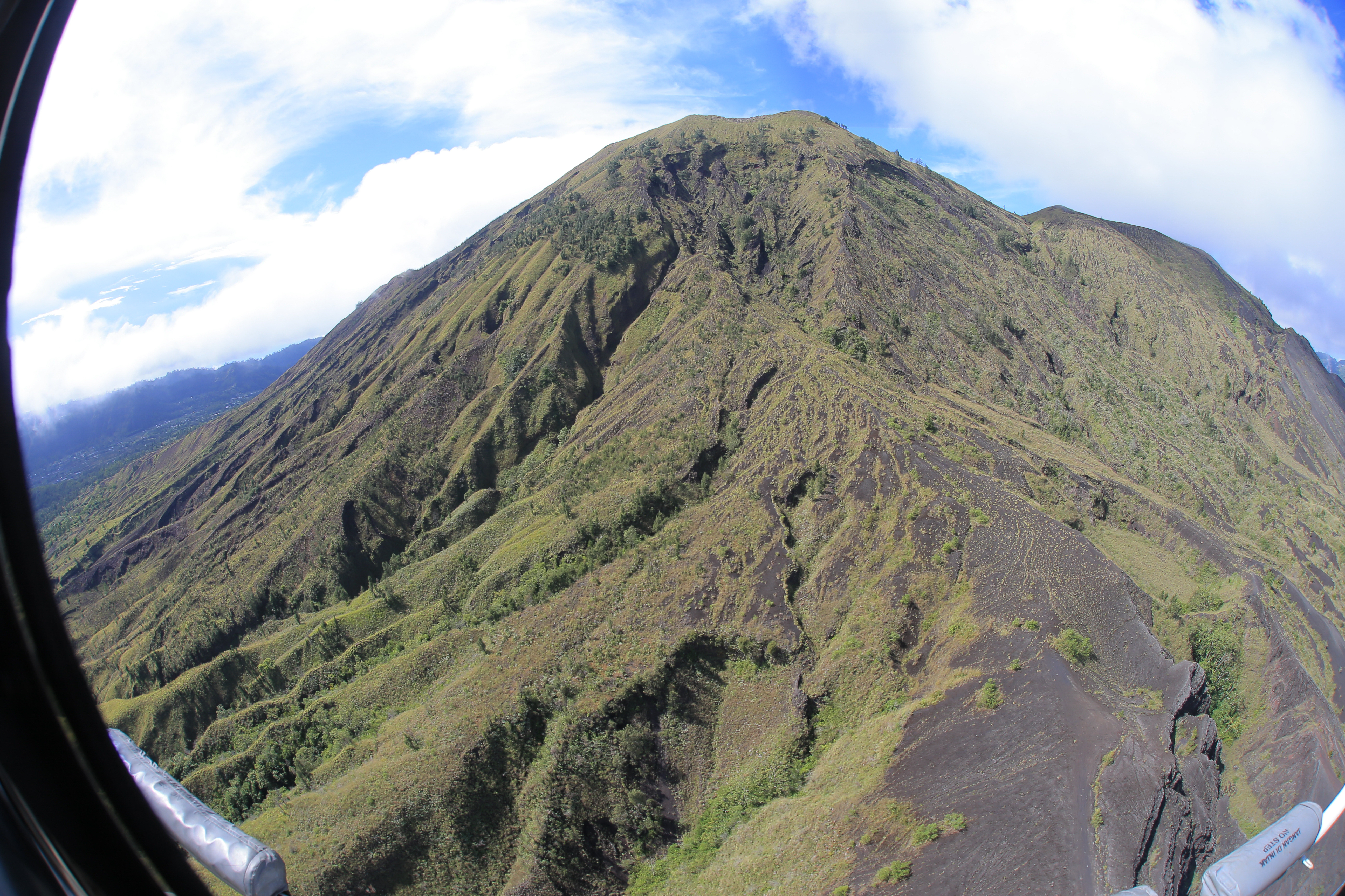 画面从飞行器舷窗视角拍摄，舷窗边缘可见“NO STEP”标识，巍峨火山铺展于蓝天白云之下，山体植被错落、沟壑纹理清晰，景致开阔壮丽，满是蓬勃向上的气息，完美呈现本次安利户外探索活动的震撼体验。