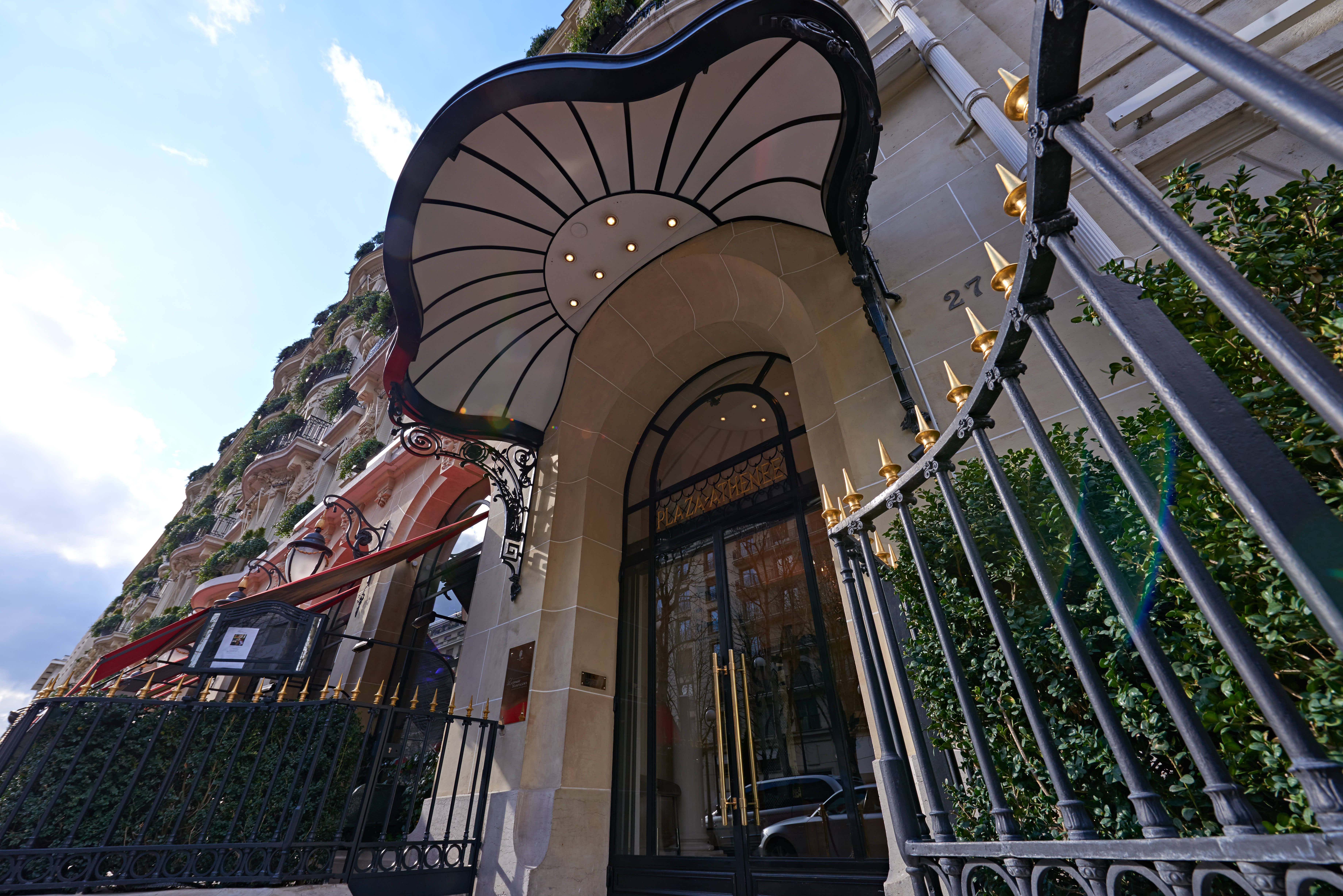 画面呈现巴黎PLAZA ATHENEE（雅典娜广场酒店）入口外景，石质建筑墙面标注门牌号27，搭配精致弧形装饰雨篷、带金色尖饰的黑金铁艺栅栏，旁侧绿植点缀，晴日下建筑格调典雅高级，是安利高端海外品牌活动的举办场地，尽显活动高端质感。