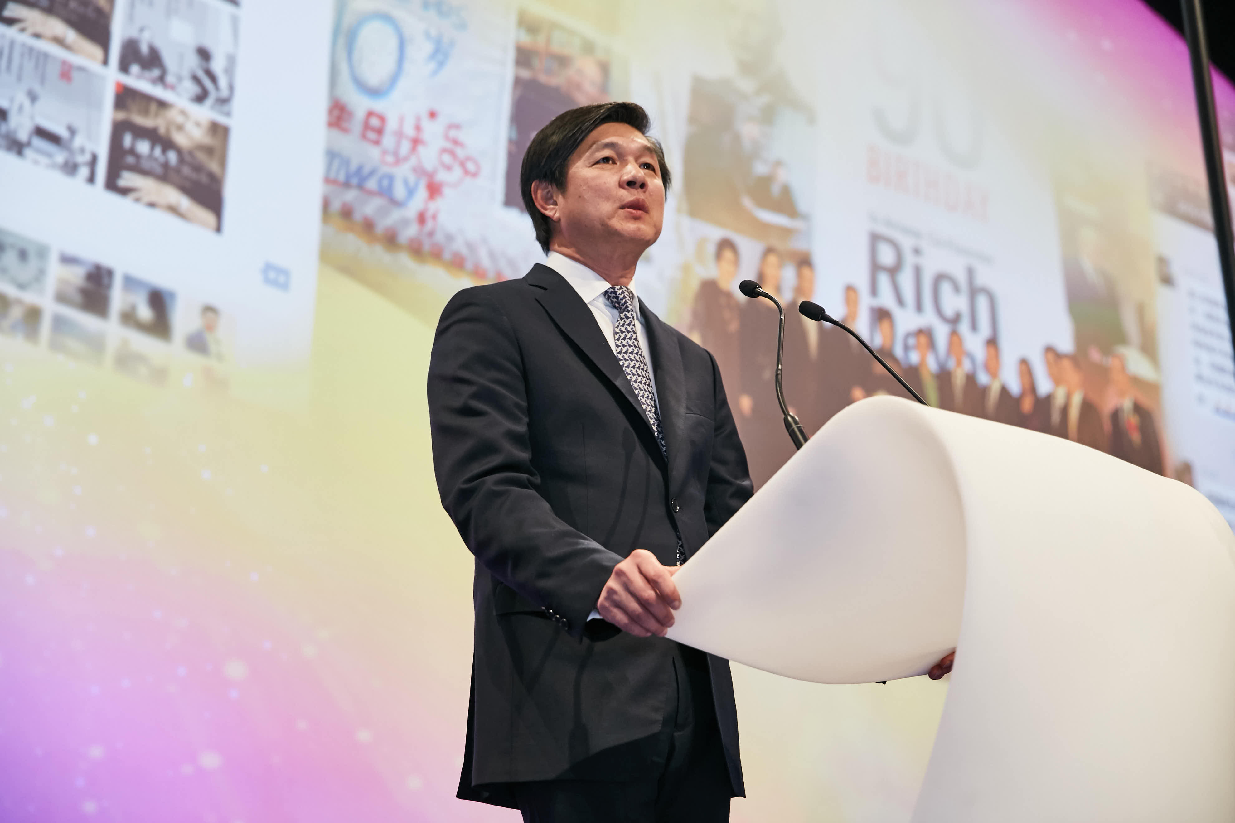 本次为安利周年庆典活动现场，身着正装的企业高管正站在白色演讲台前发言，背景大屏展示着品牌发展历程影像、“BIRTHDAY”相关字样，现场氛围庄重热烈，彰显出企业昂扬向上的品牌风貌。