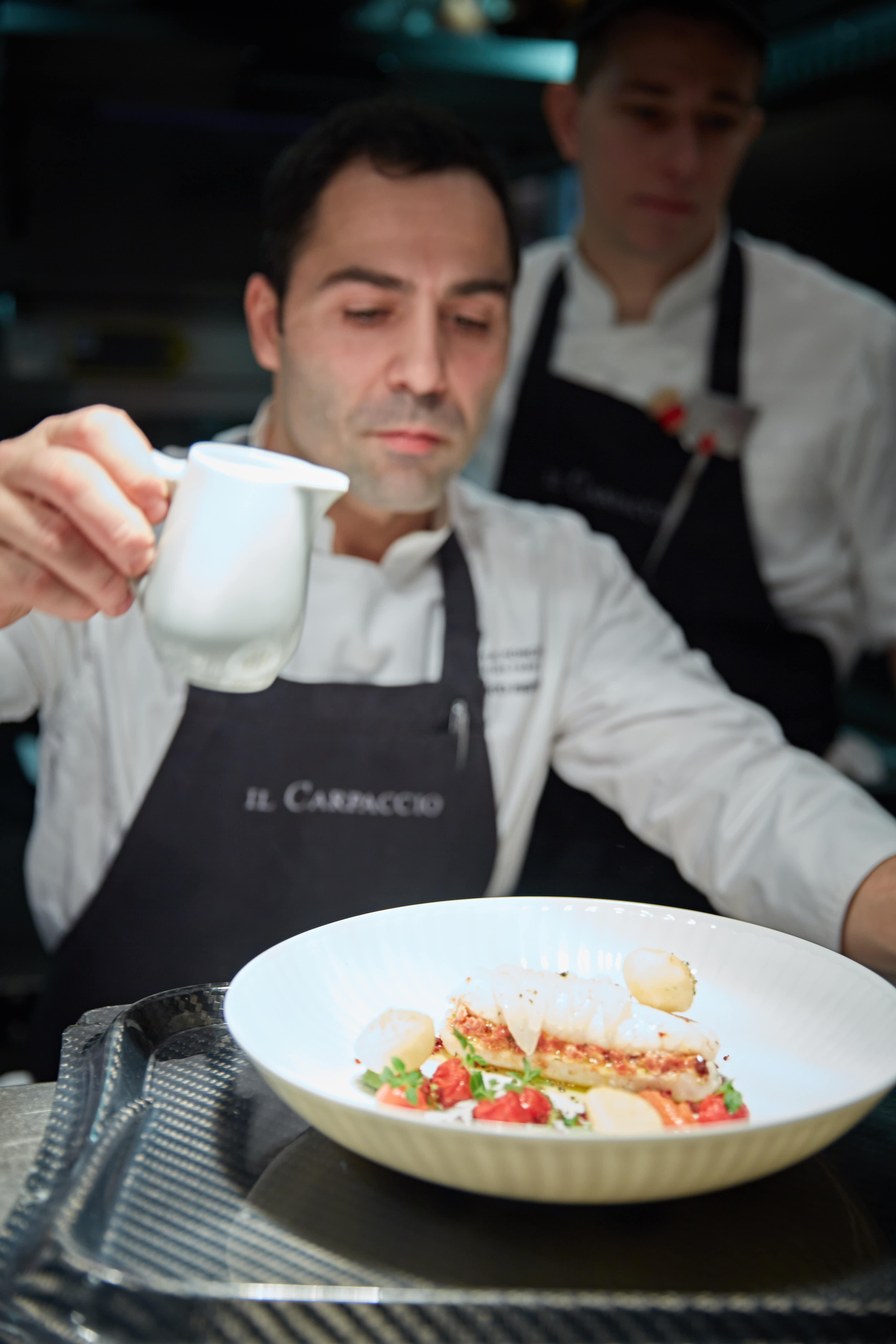安利美食体验活动现场，身着印有“IL CARPACCIO”字样围裙的外籍主厨，正专注为摆盘精致的餐品淋浇酱汁，身后另有厨师协同配合，整体氛围专业考究，尽显本次活动的高品质质感。