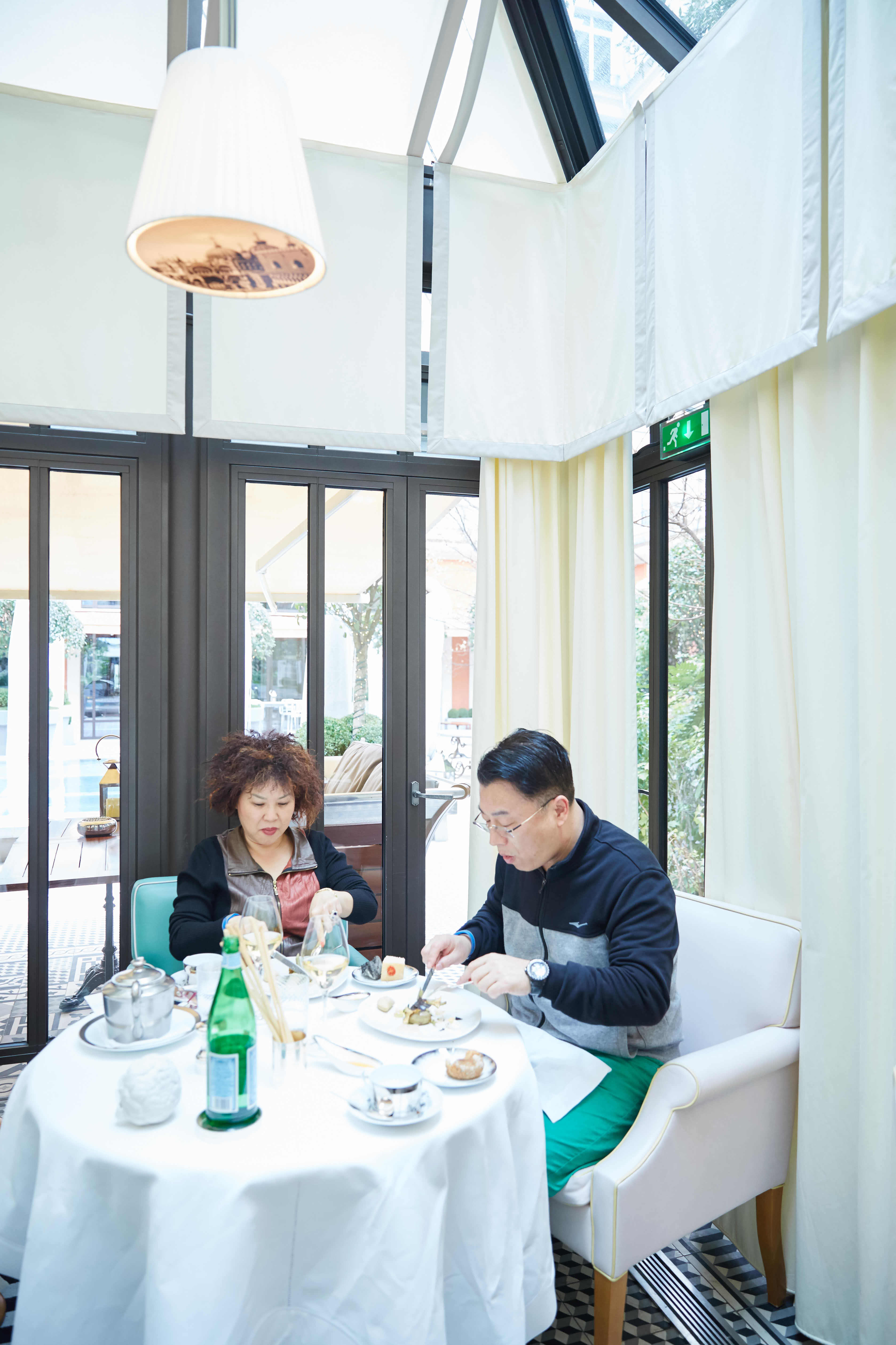 明亮通透的玻璃餐区环境雅致，两位安利伙伴围坐在铺有洁白桌布的餐桌旁，桌上摆放着精致餐食、饮品与餐具，二人正惬意享用餐点，氛围轻松闲适，展现出安利活动中伙伴们享受品质生活、舒展放松的温馨状态。
