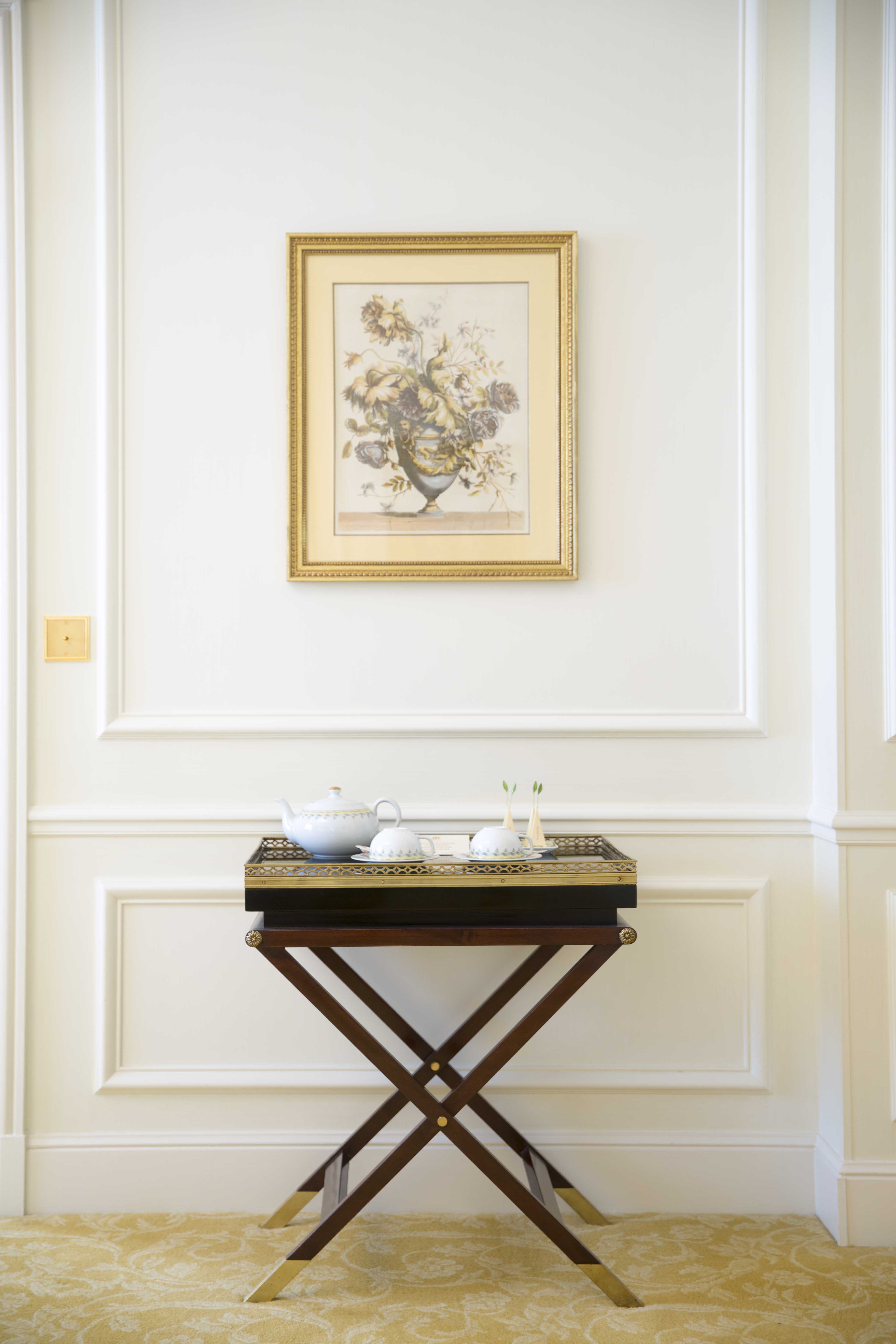 画面中米白色护墙板墙面悬挂鎏金框花卉装饰画，下方深色实木折架边几上摆放着成套白瓷茶具，整体环境雅致整洁，氛围静谧舒适，是安利活动为参会人员设置的休憩点位，尽显品牌贴心周到的服务质感。
