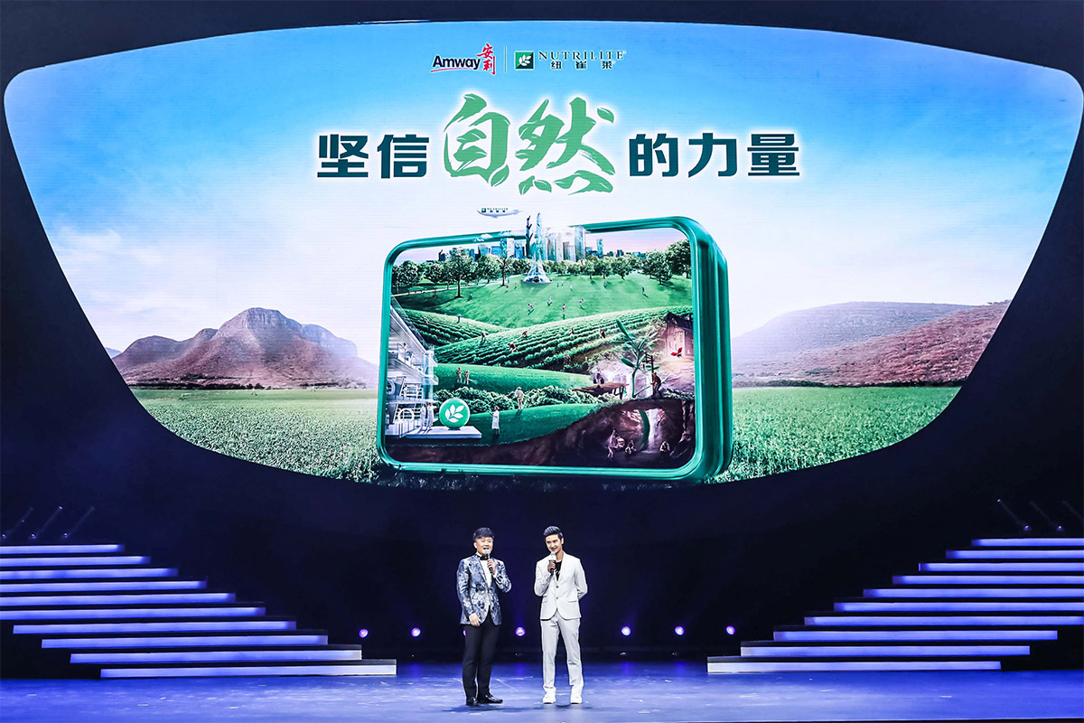 活动现场大屏展示有“Amway安利”“NUTRILITE纽崔莱”品牌标识，以及“坚信自然的力量”主题标语，大屏同步呈现自然种植场景的品牌视觉内容，两位男嘉宾持麦站在舞台上互动，现场氛围专业明快，充分传递纽崔莱依托自然的健康品牌理念。