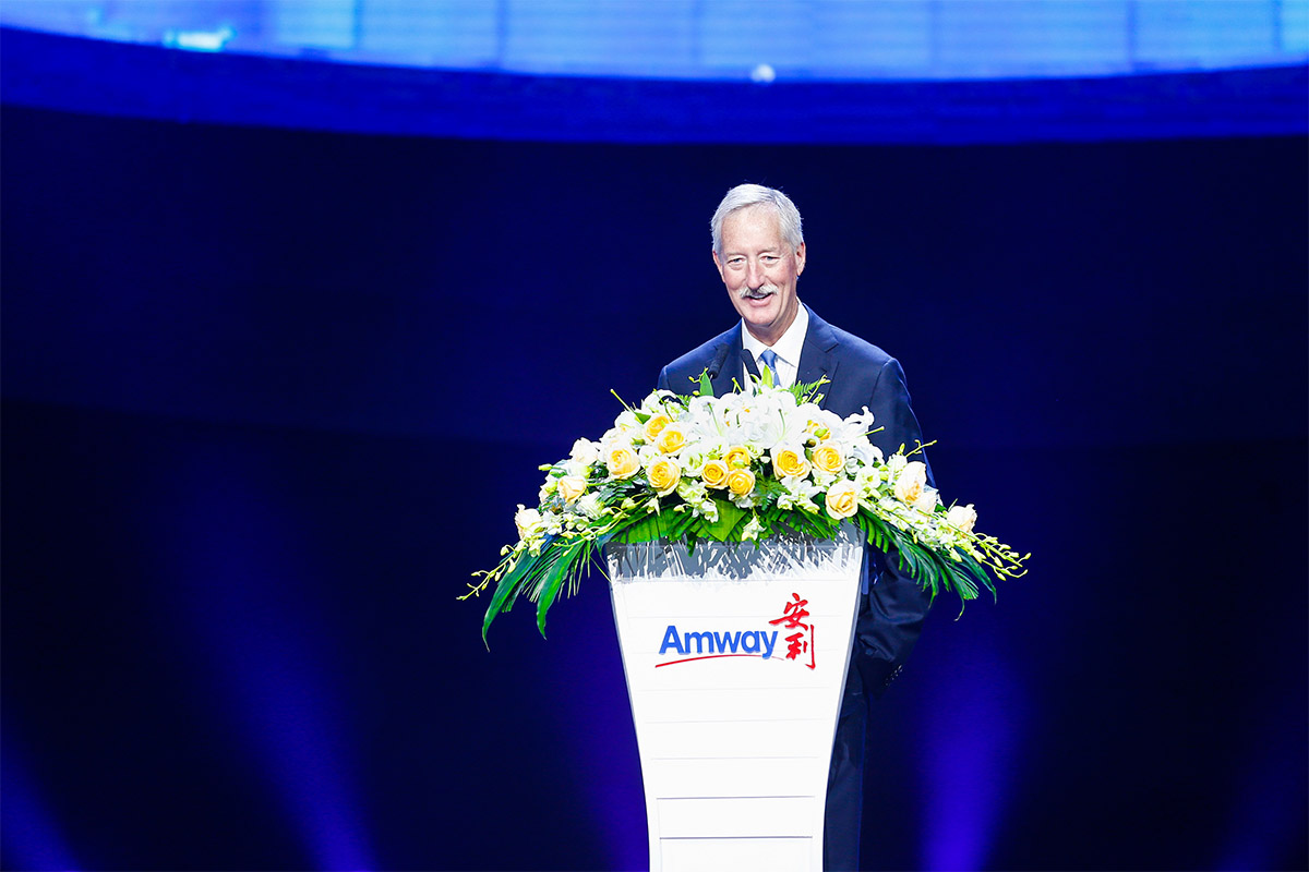 活动现场，安利外籍高管身着正装，站在印有“Amway 安利”标识的讲台后致辞。讲台上装饰有黄玫瑰、白百合等花艺，精致亮眼。蓝紫色调的舞台背景氛围感十足，高管面带笑意神情亲和，整体场景庄重正向，契合品牌活动的正式调性。