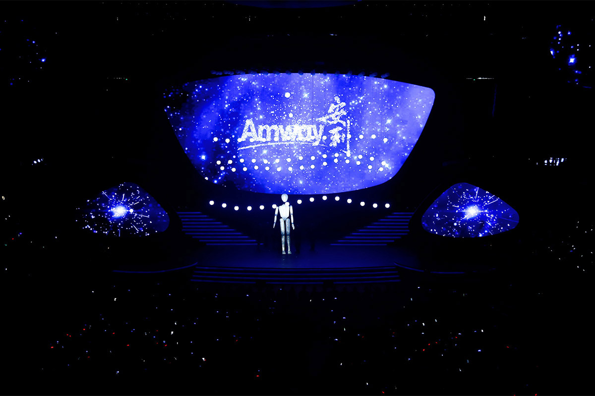 活动现场主屏幕以浩瀚星空为视觉基底，醒目展示“Amway 安利”品牌标识，两侧副屏呼应星芒特效，舞台中央设立人形机器人装置，整体蓝调光影充满未来科技感，台下观众席星光点点，氛围隆重且富有创意，彰显安利的创新品牌调性。