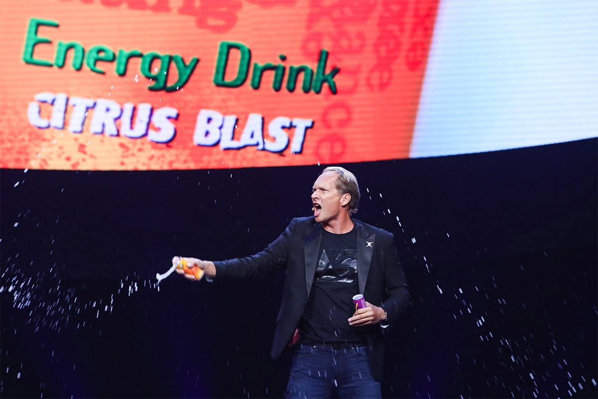 安利活动现场背景大屏醒目显示“Energy Drink”“CITRUS BLAST”字样，主讲人身着带有XS标识的服饰，手持两款XS能量饮料，正向现场喷洒饮品，飞溅的液滴充满动感，现场氛围热烈鲜活，充分传递XS能量饮的活力属性。