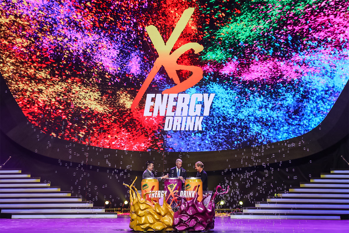 活动现场背景大屏呈现炫彩粒子效果，醒目展示“XS ENERGY DRINK”标识。三位嘉宾立于XS能量饮料造型的创意启动装置旁共同完成启动环节，现场飘满透明气泡，氛围热烈鲜活，充分彰显XS品牌年轻动感的特质，传递满满活力。