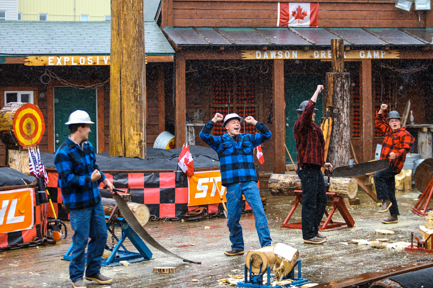 本次活动在道森溪营地（DAWSON CREEK CAMP）开展，细雨中，参与伐木体验的安利伙伴身着格纹工装、佩戴安全帽，完成比拼后高举手臂欢呼，周边摆放着锯木工具与原木，现场氛围热烈昂扬，尽显伙伴们的活力与拼搏劲头。