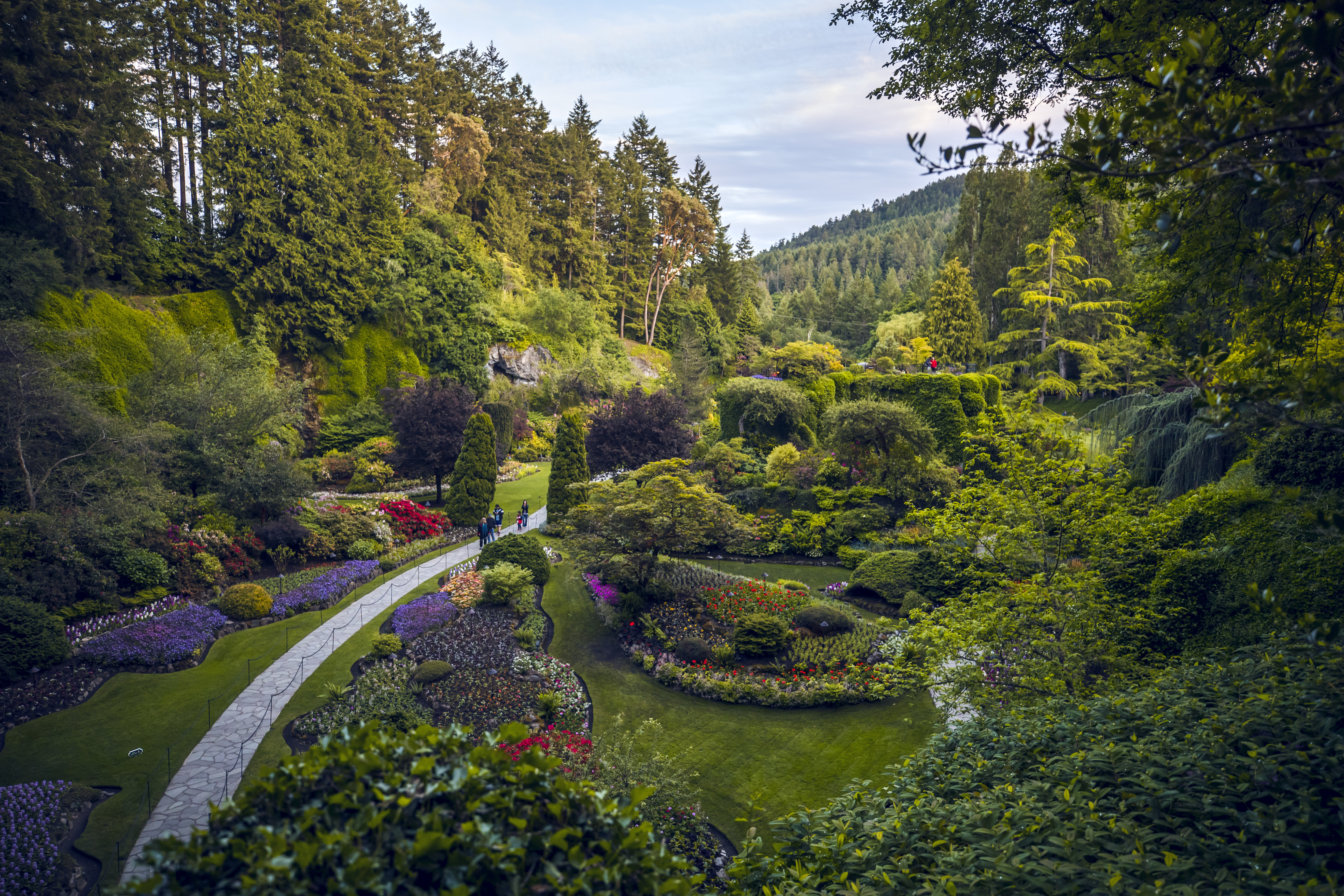 画面呈现了植被繁茂、花团错落的清幽园林盛景，石质步道上有结伴游览的人员，整体氛围清新自然、生机盎然，契合安利健康自然的品牌调性，尽显团队户外出行的轻松愉悦氛围。