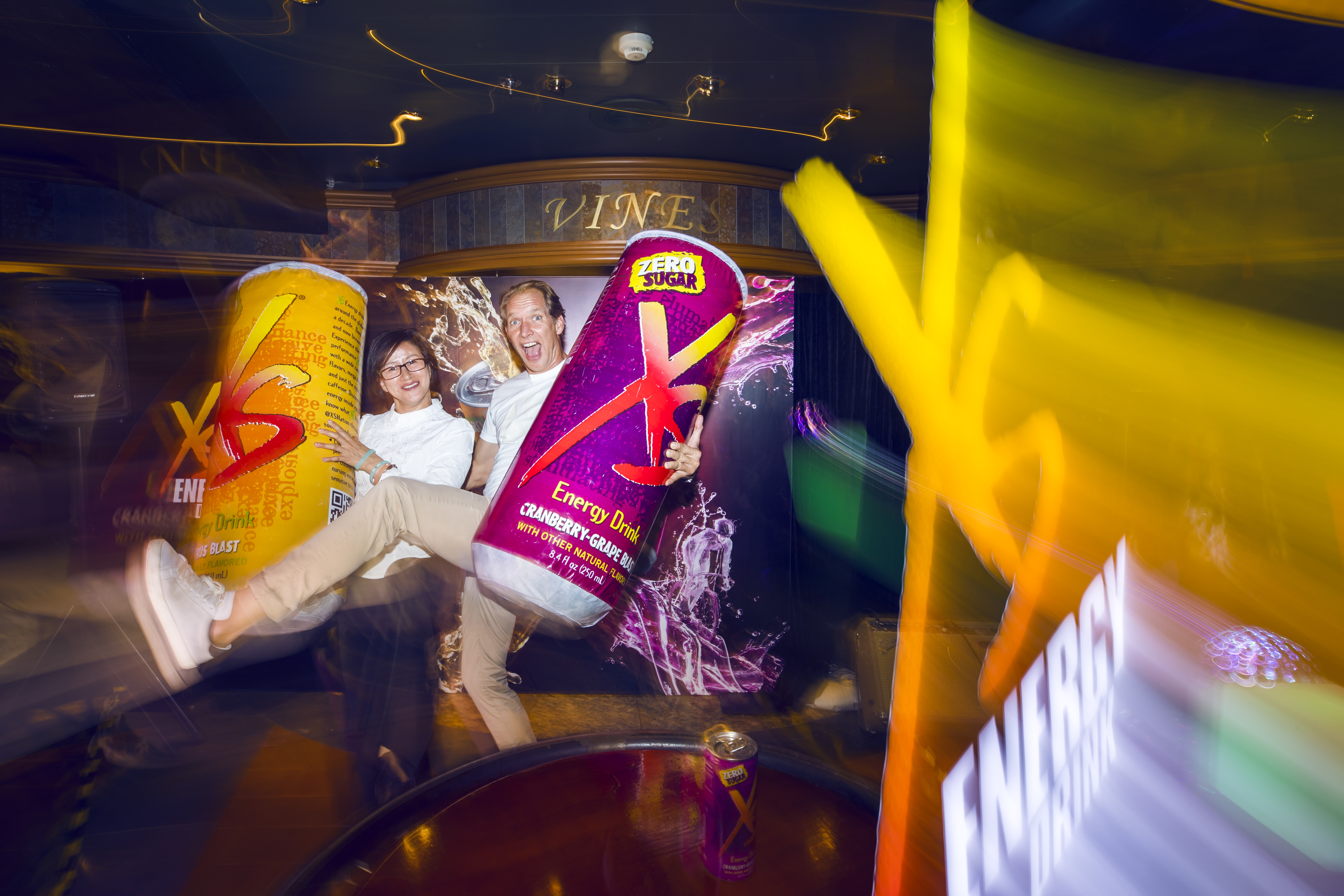 动感光影的活动现场中，两位参与者手持印有“ZERO SUGAR”“Energy Drink”字样的XS能量饮料巨型充气模型，表情愉悦动作活泼，背景搭配品牌宣传海报，整体氛围活力热烈，尽显安利XS能量饮年轻动感的品牌特质。