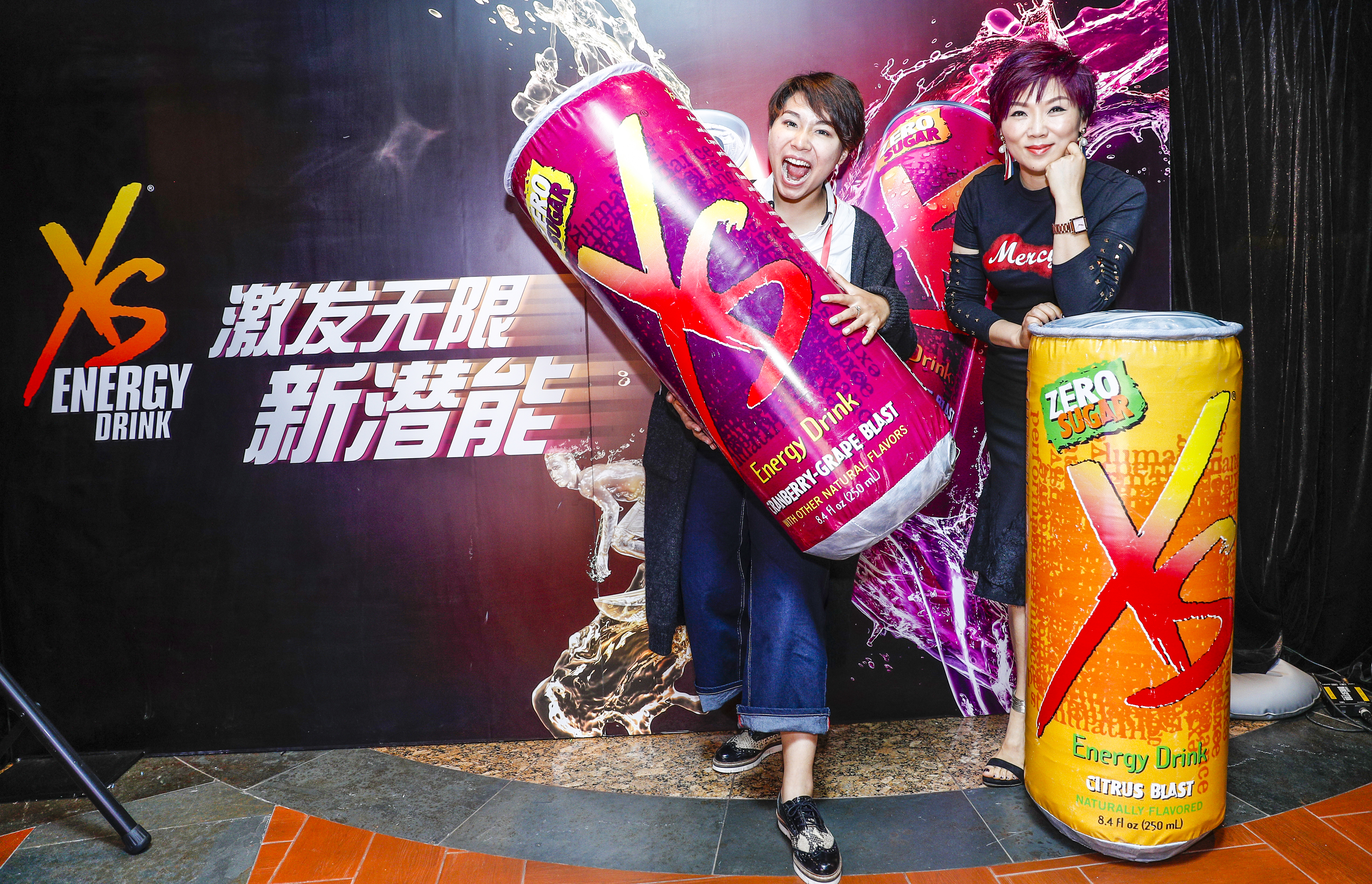 背景印有“XS ENERGY DRINK”“激发无限新潜能”字样，两位参与者在活动现场打卡，一位怀抱巨型蔓越莓葡萄味XS能量饮料气模，另一位倚靠柑橘味零糖XS能量饮料气模，现场氛围鲜活热烈，尽显XS品牌活力向上的特质，传递迸发潜能的积极理念。