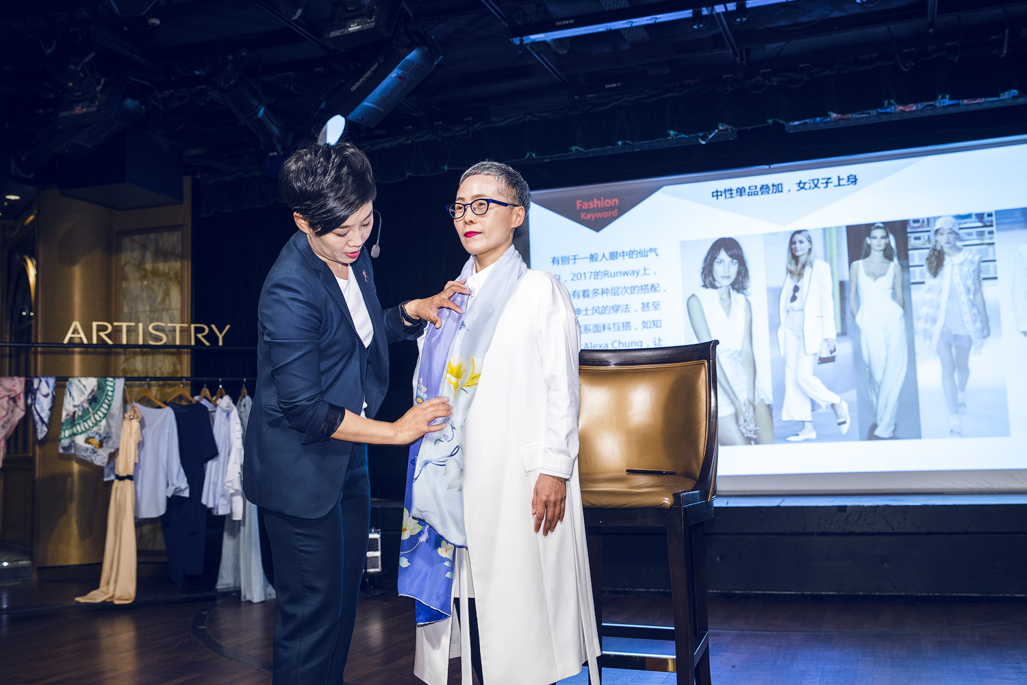 本次为安利雅姿（ARTISTRY）时尚穿搭分享活动现场，专业讲师正为体验者调整丝巾搭配造型，身后大屏展示时尚穿搭关键词及穿搭参考案例，现场还陈列有多款服饰样品，氛围专业轻松，向参与者传递实用穿搭技巧与时尚美学生活理念。
