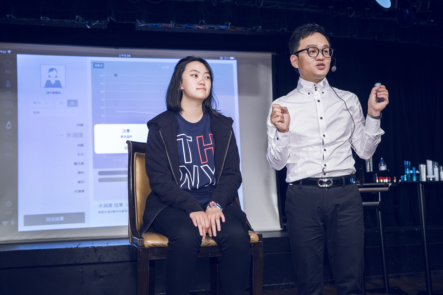 活动现场讲师佩戴耳麦手持护肤样品讲解，体验者落座配合演示，身后投影屏幕展示肤质检测界面，包含水润度、测试结果等相关数据，侧台摆放多款安利护肤产品，现场氛围专业务实，充分展现安利科学护肤的产品理念。