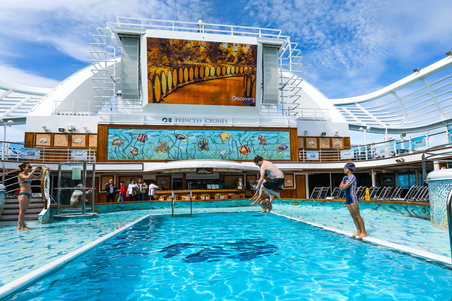 本次安利专属旅程搭载公主邮轮（PRINCESS CRUISES），船顶大屏显示“Discovery AT SEA”字样。画面展现邮轮甲板泳池区的欢乐场景，蓝天白云下澄澈的泳池边，伙伴们或跃入水中嬉戏，或拍照记录美好瞬间，氛围轻松愉悦，尽显旅程的惬意舒适与鲜活活力。