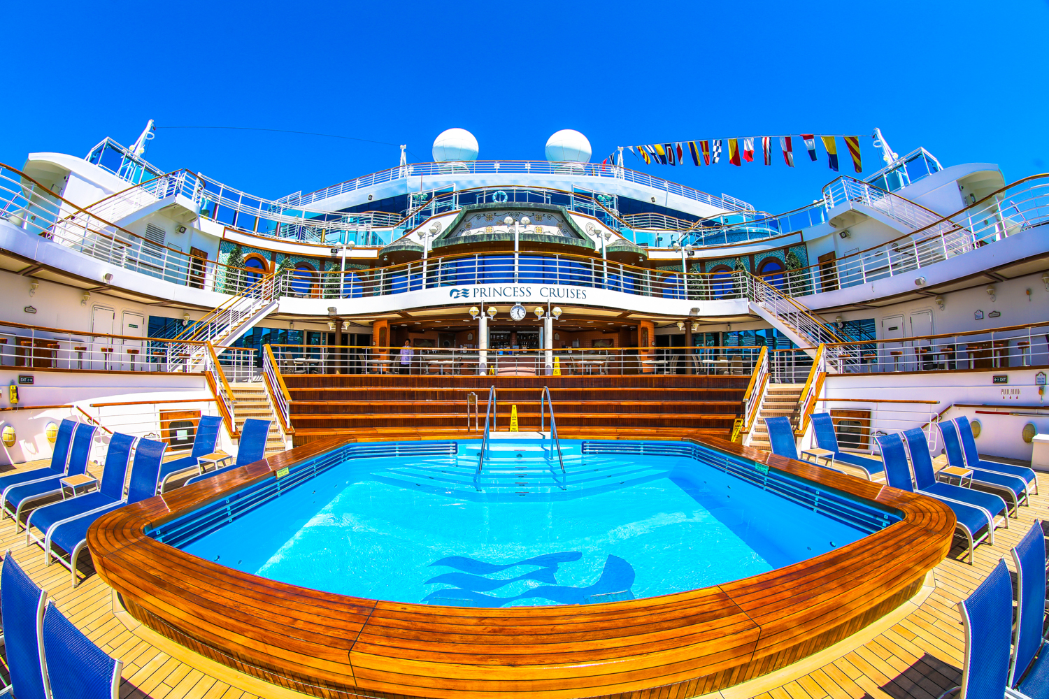 画面为公主邮轮（PRINCESS CRUISES）露天泳池区域，澄澈泳池搭配木质包边，周边整齐排布休闲躺椅，邮轮上层建筑整洁大气，晴空湛蓝明朗，整体氛围舒适松弛，是安利为参与活动的伙伴打造的高端休闲场景，尽显品牌贴心福利与高端调性。