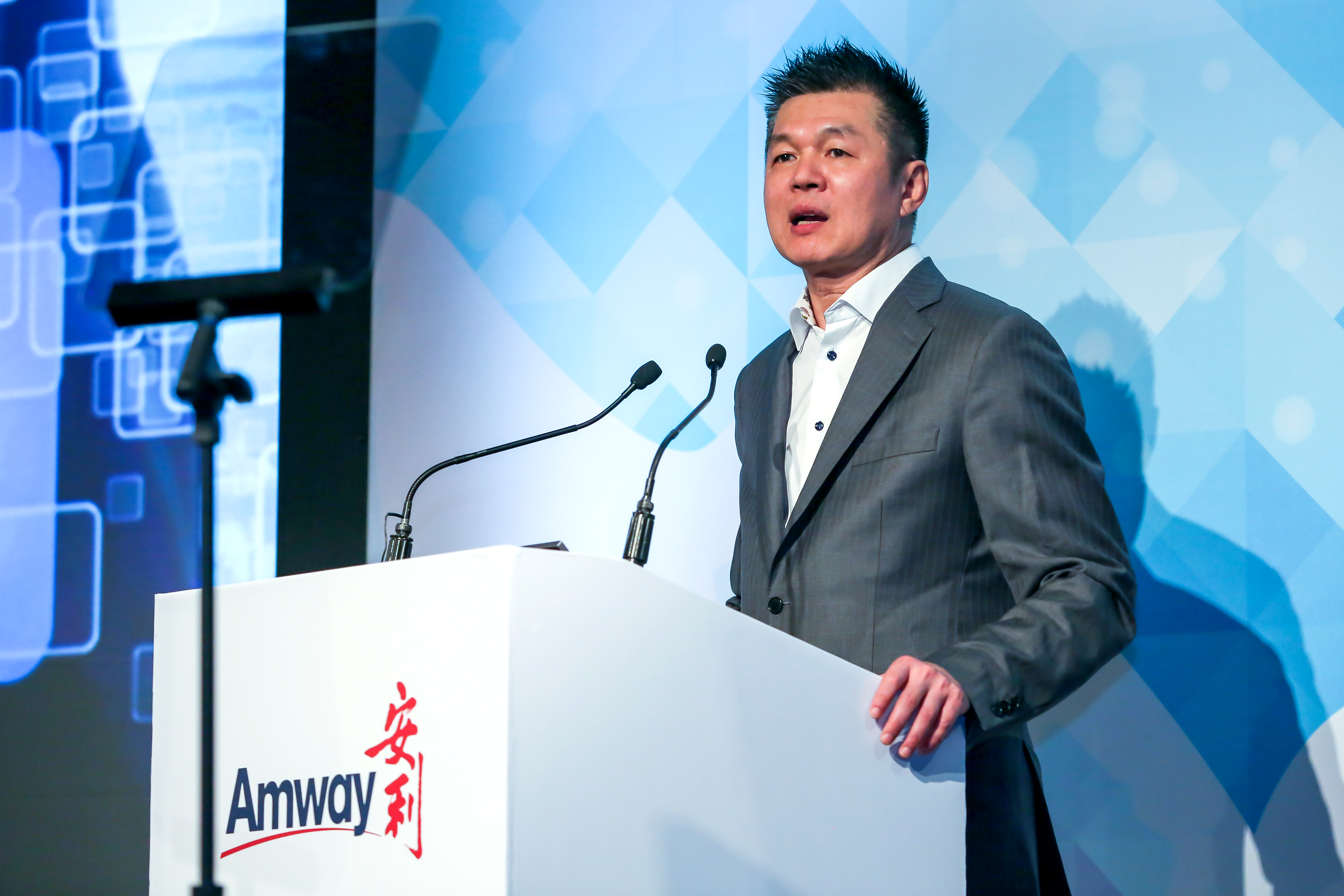 活动现场，身着灰色西装的男士站在印有“Amway 安利”标识的白色演讲台后发言，演讲台配备专业拾音话筒，背景为蓝调科技感几何大屏，整体氛围正式专业，彰显安利品牌活动的专业质感与正向风貌。