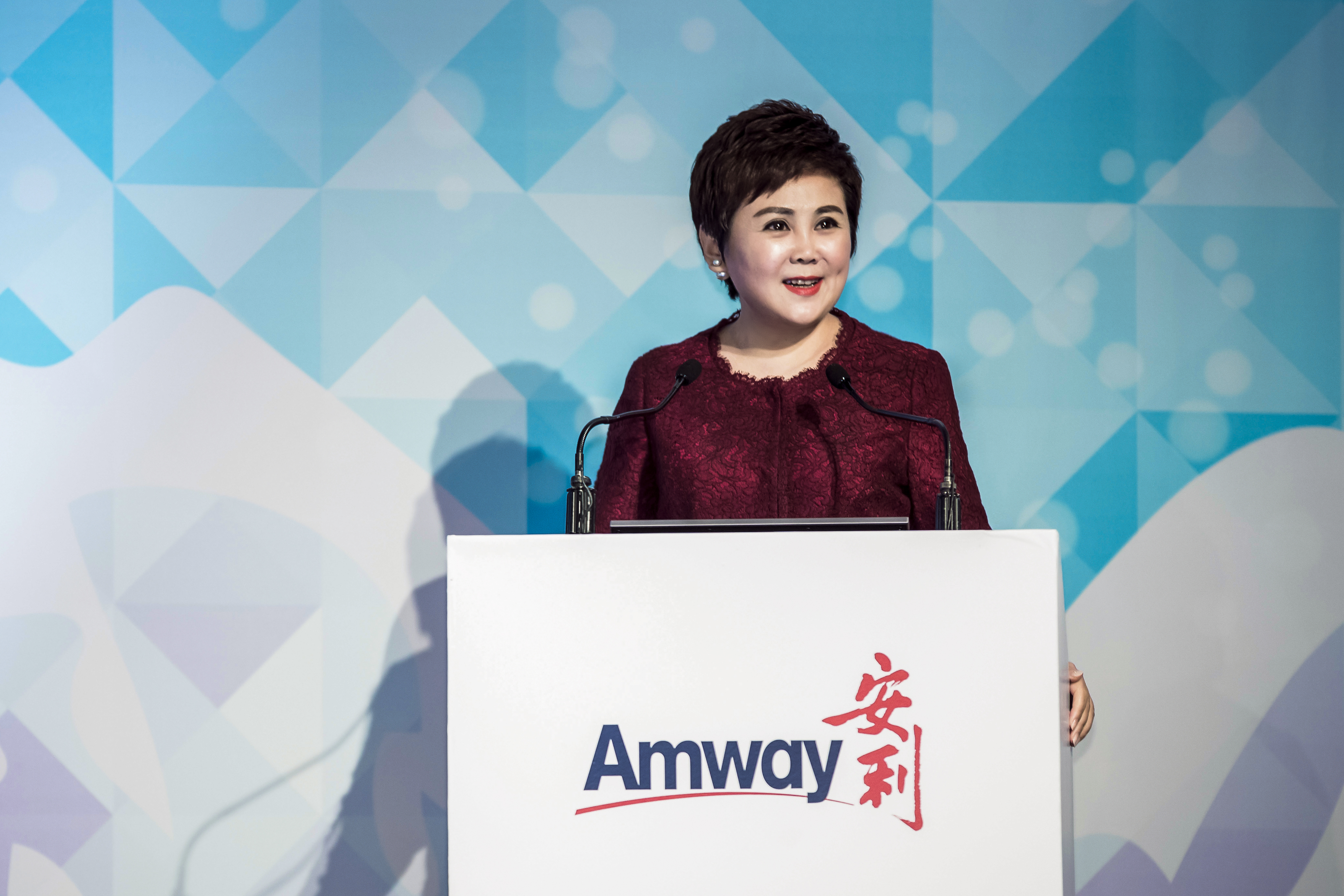 活动现场，身着酒红色上衣的发言嘉宾站在印有“Amway 安利”标识的白色演讲台后致辞，背景为蓝白几何纹理搭配光斑的设计背板，嘉宾神态亲和从容，现场氛围正式积极，尽显安利品牌活动的专业正向风貌。