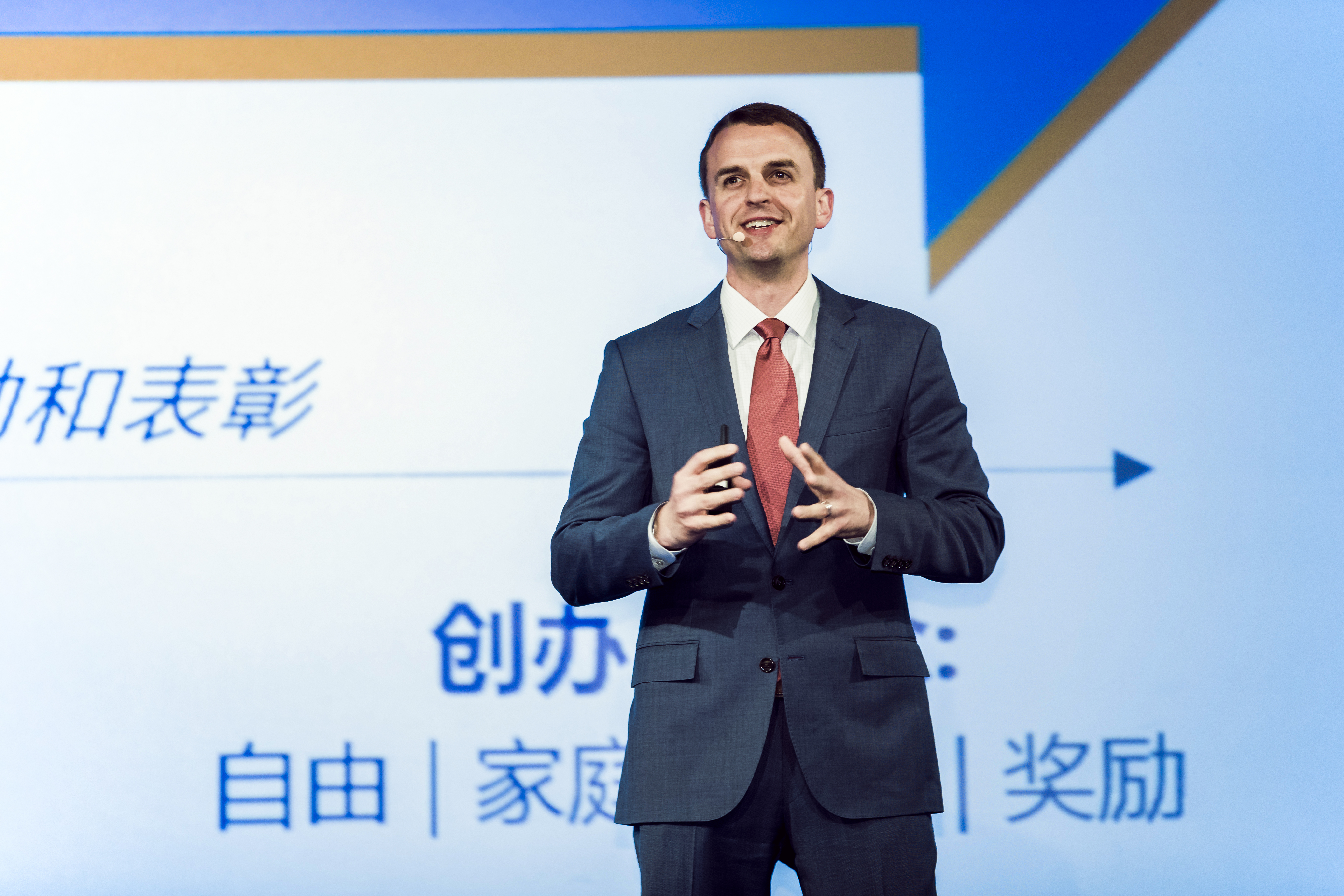 安利品牌活动现场，身着正装的外籍高管佩戴耳麦、手持演示器，正在台上进行主题分享。身后大屏显示“力和表彰”“创办”“自由 | 家庭 | 奖励”等字样，现场氛围专业积极，清晰传递品牌发展理念，为参会伙伴明晰发展方向。