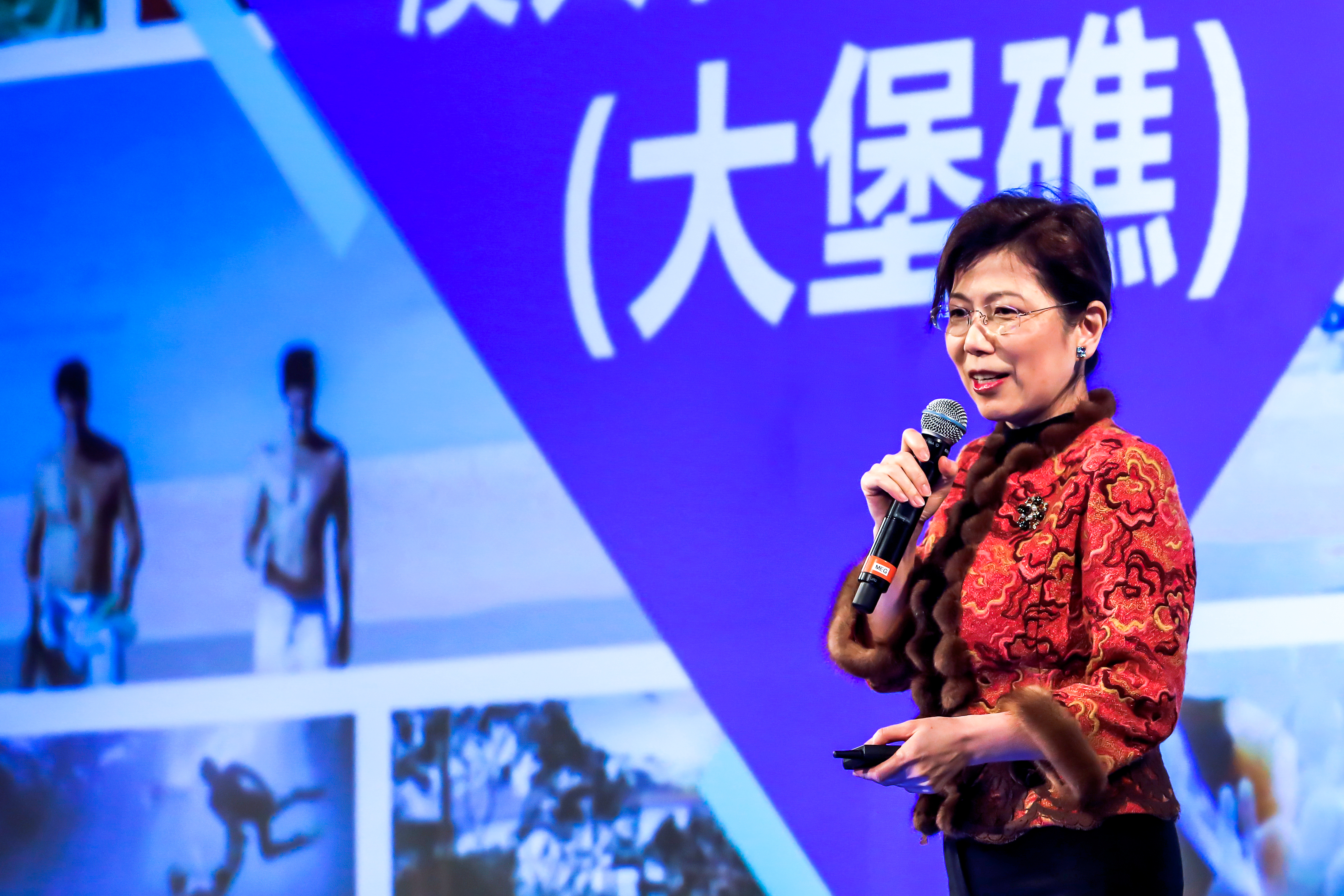本次安利品牌活动现场，身着精致中式红装的女性嘉宾手持麦克风与翻页器正在分享，背景大屏清晰显示核心文字“大堡礁”及相关场景影像，现场氛围专业热烈，充分彰显安利积极向上的活动调性。