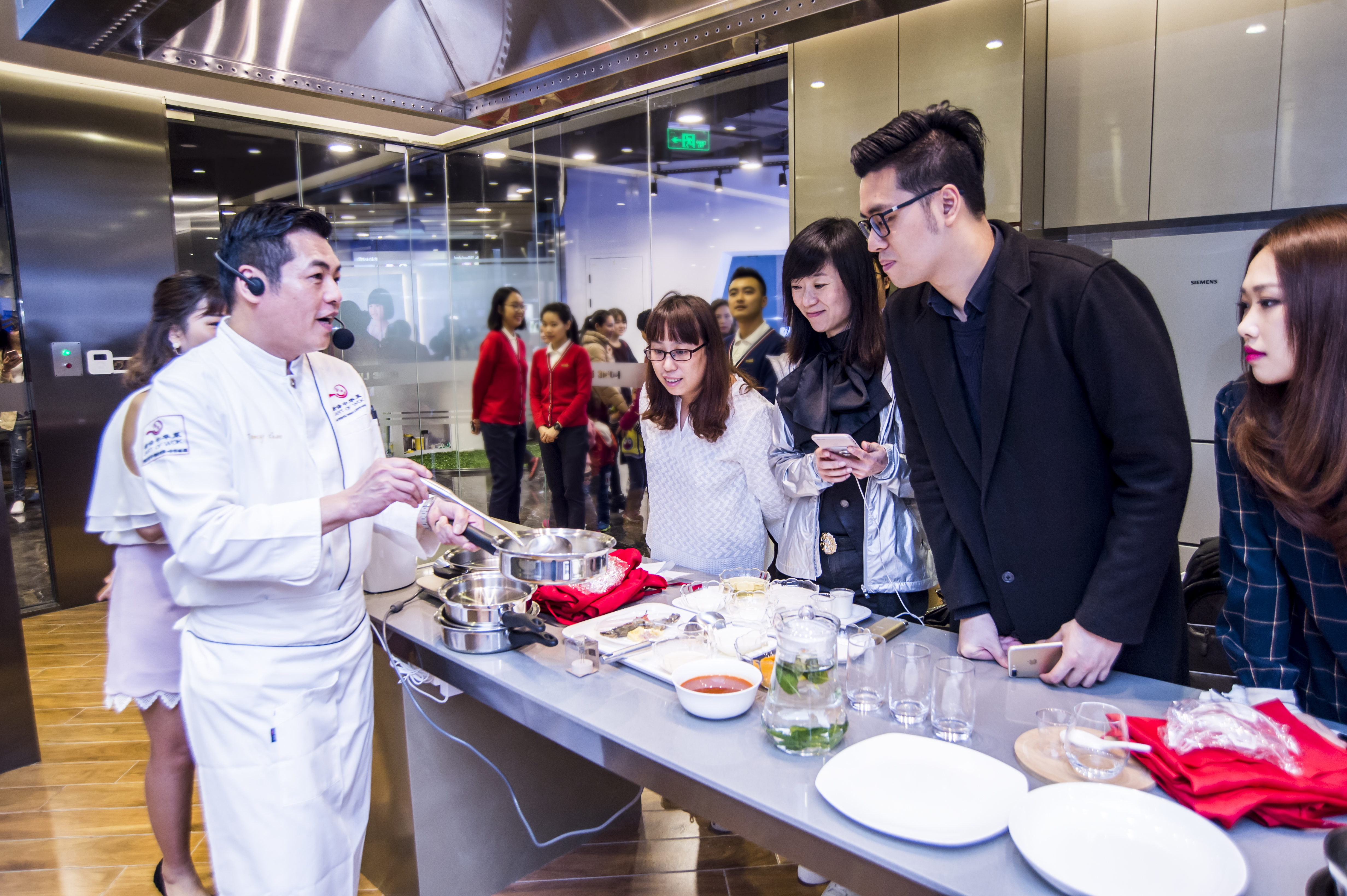 本次安利美食体验活动现场，专业厨师佩戴耳麦在操作台前，为参与活动的伙伴演示烹饪技巧，台面上摆放着锅具、食材、餐具等物料，在场参与者围立在桌旁专注聆听观看，氛围轻松活跃，充分传递出安利倡导的健康生活理念。