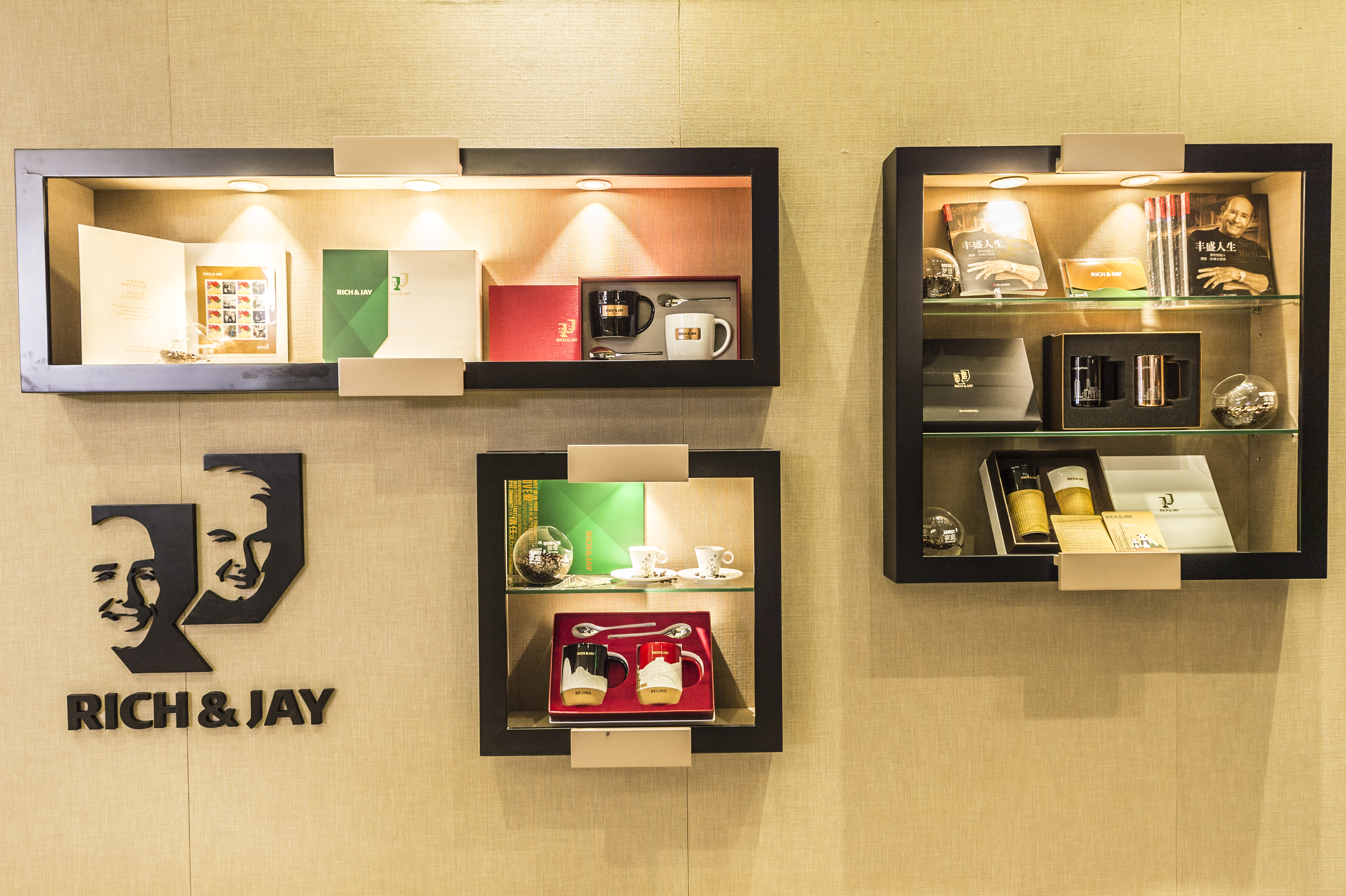 墙面印有RICH&JAY创始人标识，三处带照明的嵌入式展柜有序排布，柜内陈列《丰盛人生》书籍、品牌文创杯具、周边礼盒等物料，整体质感精良、布局规整，充分展现安利品牌文化积淀，传递品牌理念。