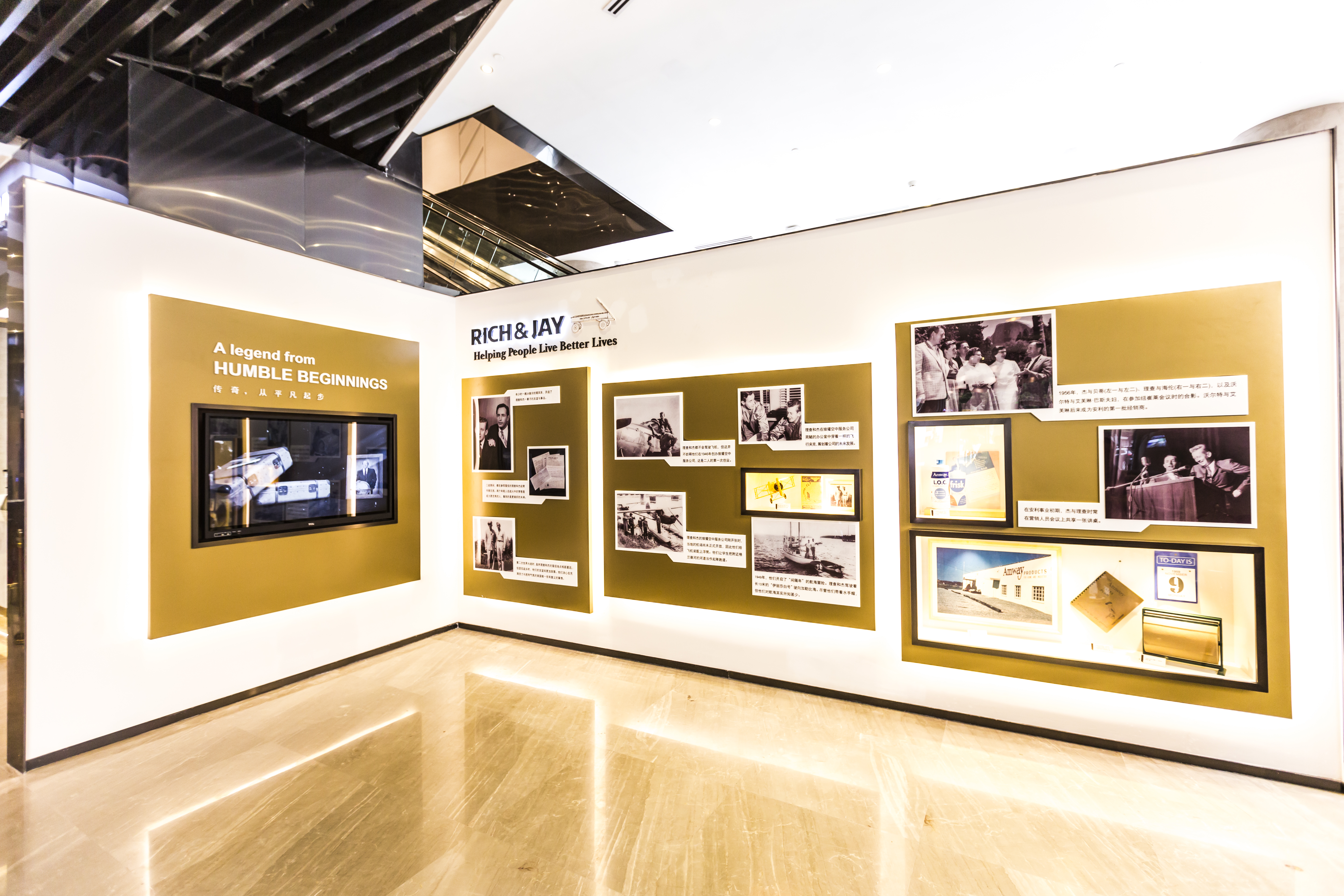 展示墙标注“RICH&JAY Helping People Live Better Lives”、“A legend from HUMBLE BEGINNINGS 传奇，从平凡起步”字样，陈列品牌发展相关老照片、历史史料，搭配宣传显示屏，整体氛围庄重明亮，清晰梳理安利发展脉络，展现品牌从平凡起步的成长历程。