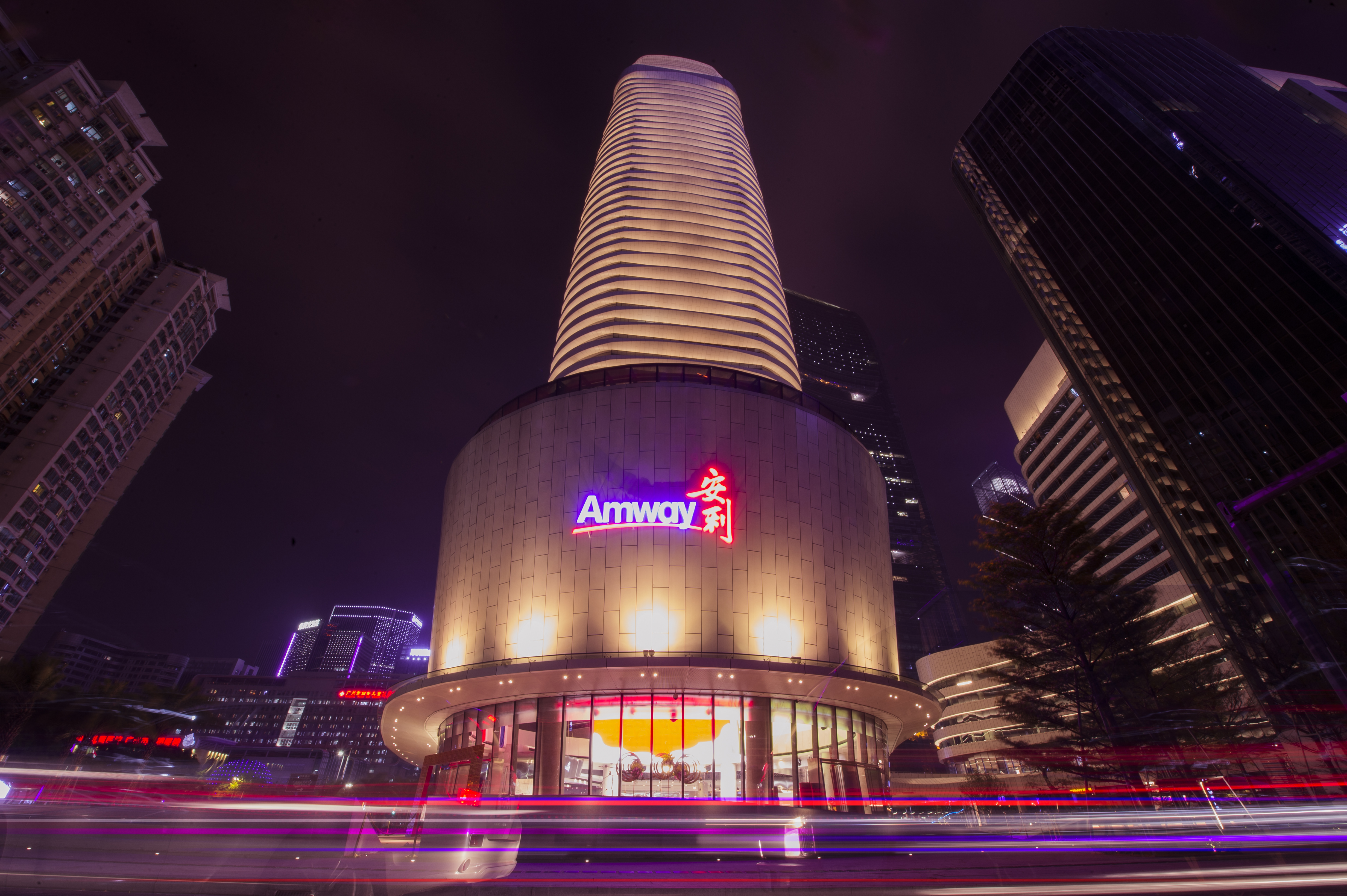 夜幕下的城市CBD核心区域，安利大楼外立面亮着醒目的“Amway 安利”品牌标识，楼体暖照明朗柔和，底层场馆通透敞亮，周边摩天楼宇林立，路面流动的车流光轨添了动感，整体氛围繁华大气，尽显安利品牌的硬核实力与城市影响力。