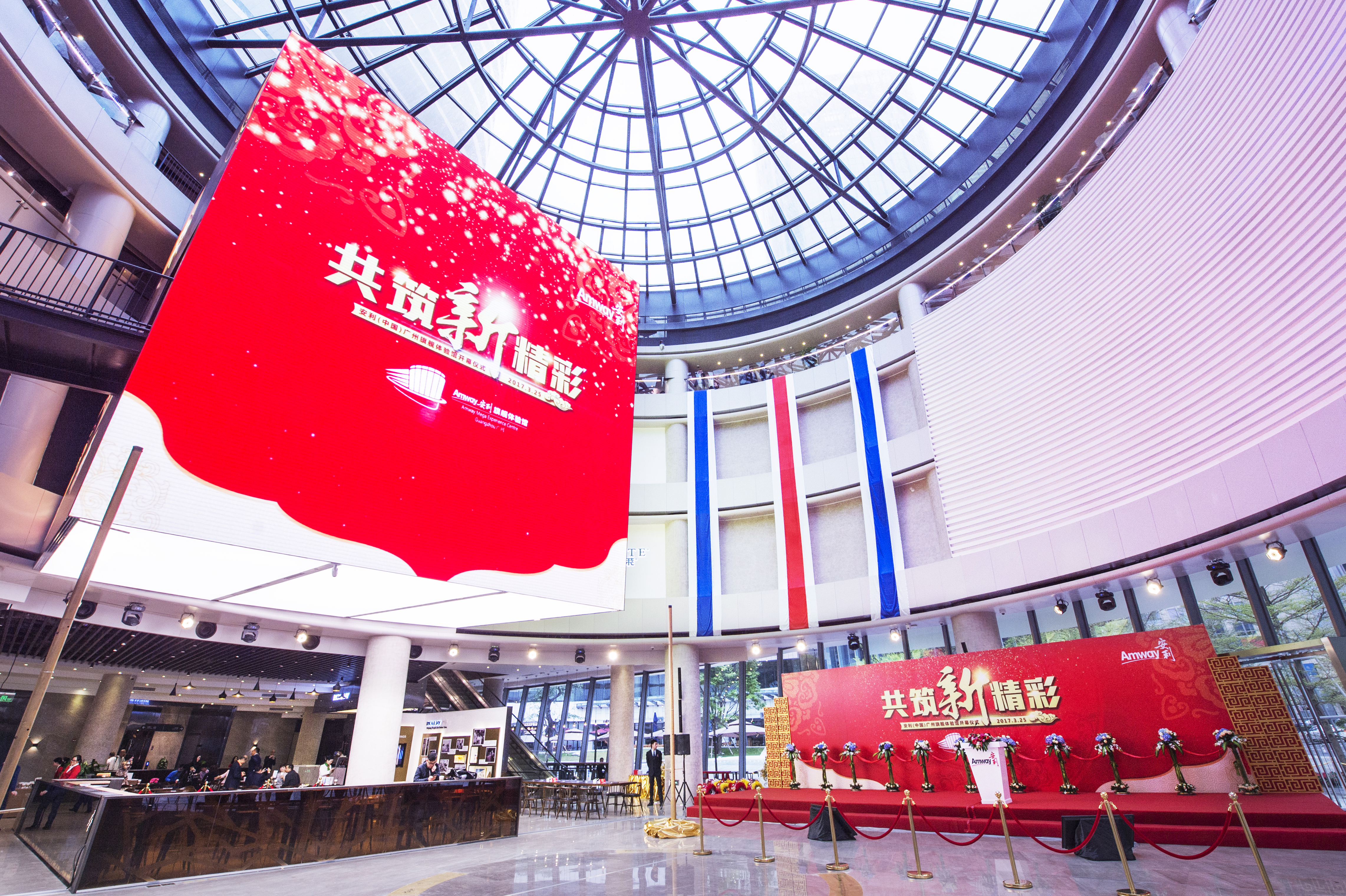画面为安利（中国）广州旗舰体验馆开馆仪式现场，挑高玻璃穹顶大厅通透明亮，两块红色主视觉屏醒目展示“共筑新精彩”活动主题，下方布置规整的红色仪式舞台，整体氛围隆重正式，尽显品牌积极向上的调性。