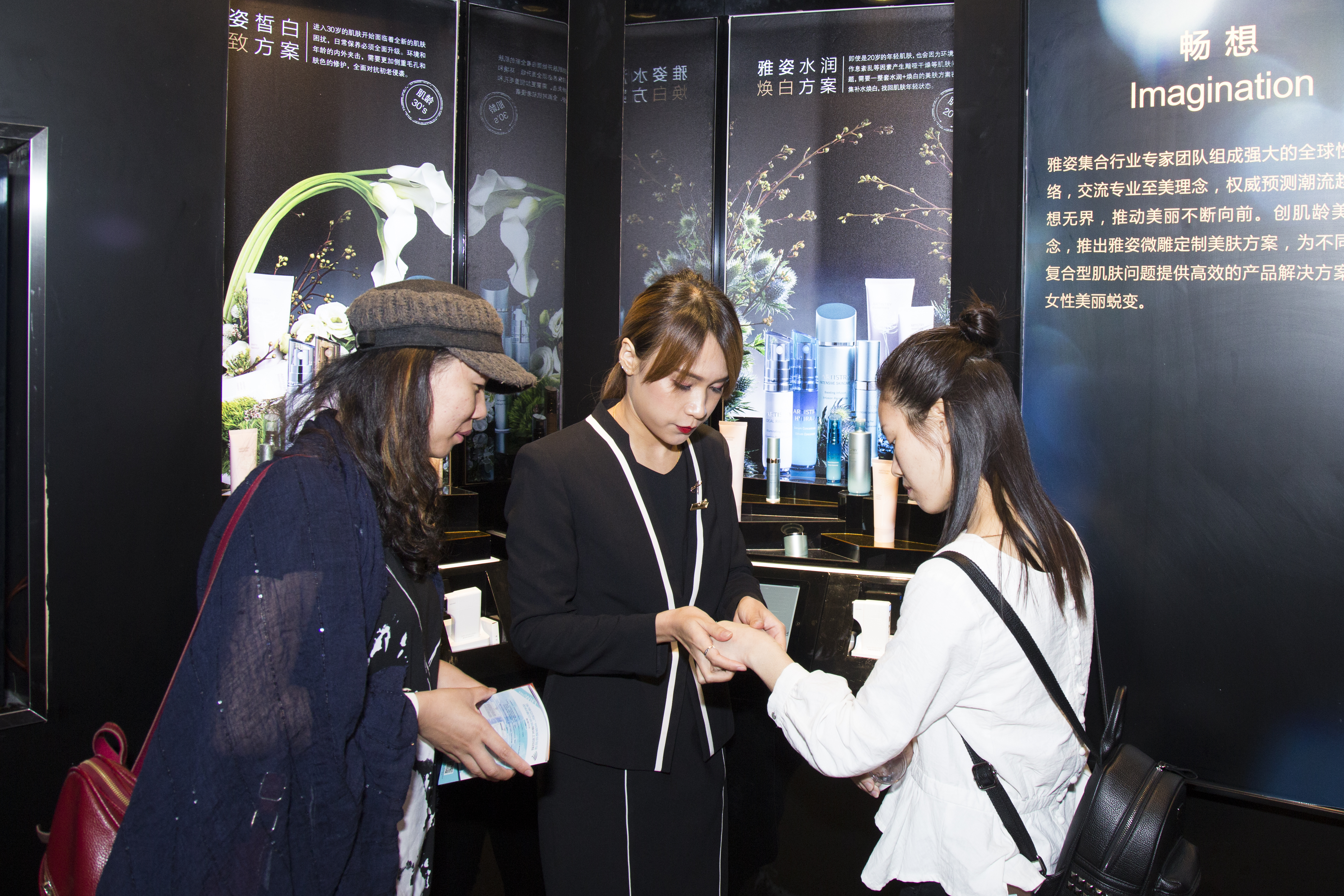 活动现场背景展板展示雅姿水润焕白方案、雅姿微雕定制美肤方案相关内容，专业工作人员正为到场体验者试用雅姿护肤产品，一旁参与者认真了解相关产品信息，现场氛围专业亲和，充分传递雅姿专业美肤的品牌理念，为消费者带来直观的护肤体验。