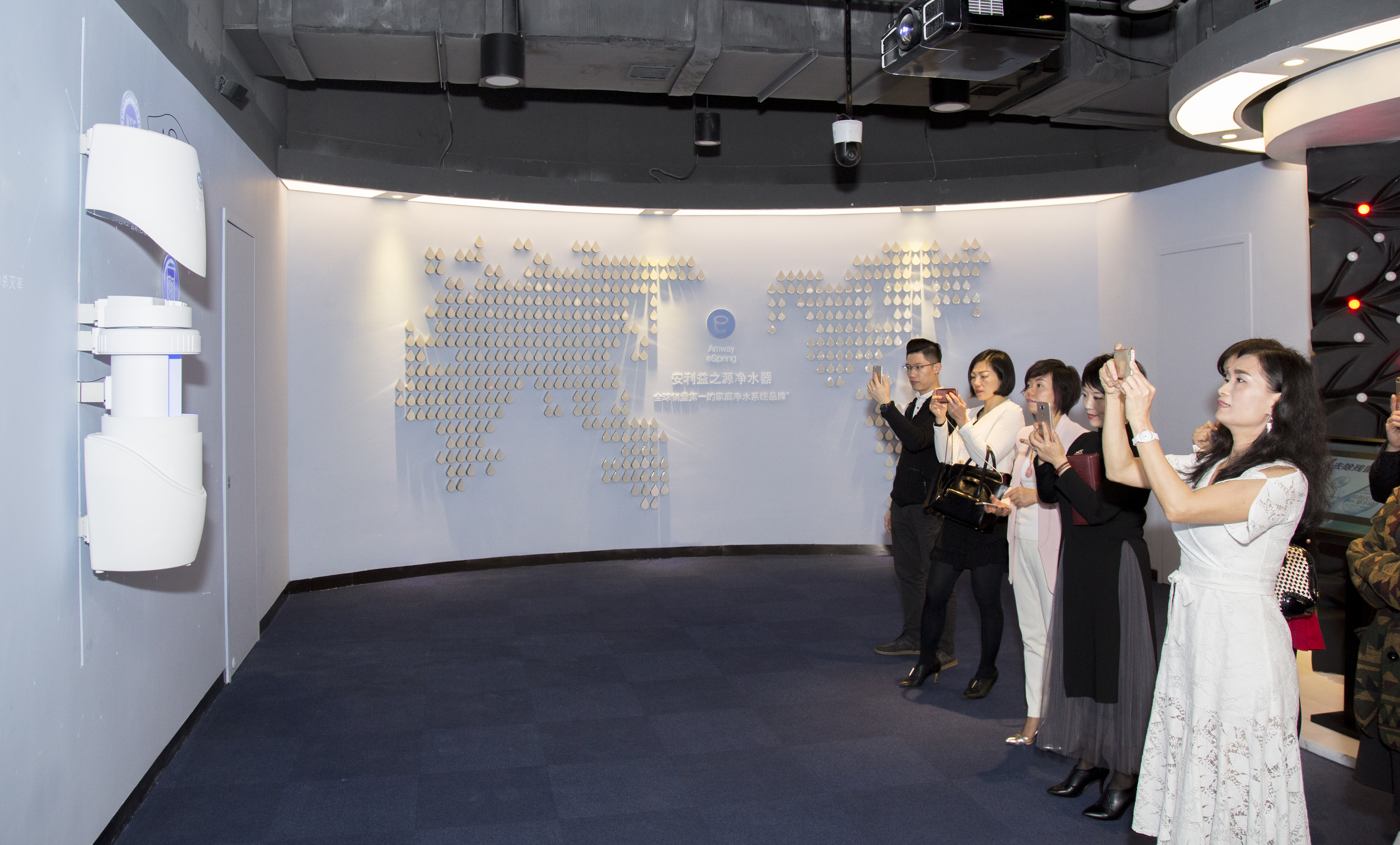 背景展示墙标注“安利益之源净水器 全球销量第一的家庭净水系统品牌”，墙面以水滴元素组成世界地图造型，左侧陈列着益之源净水器产品。多名安利合作伙伴正举手机拍摄记录，现场氛围积极专注，展现出大家对品牌产品的高度关注。
