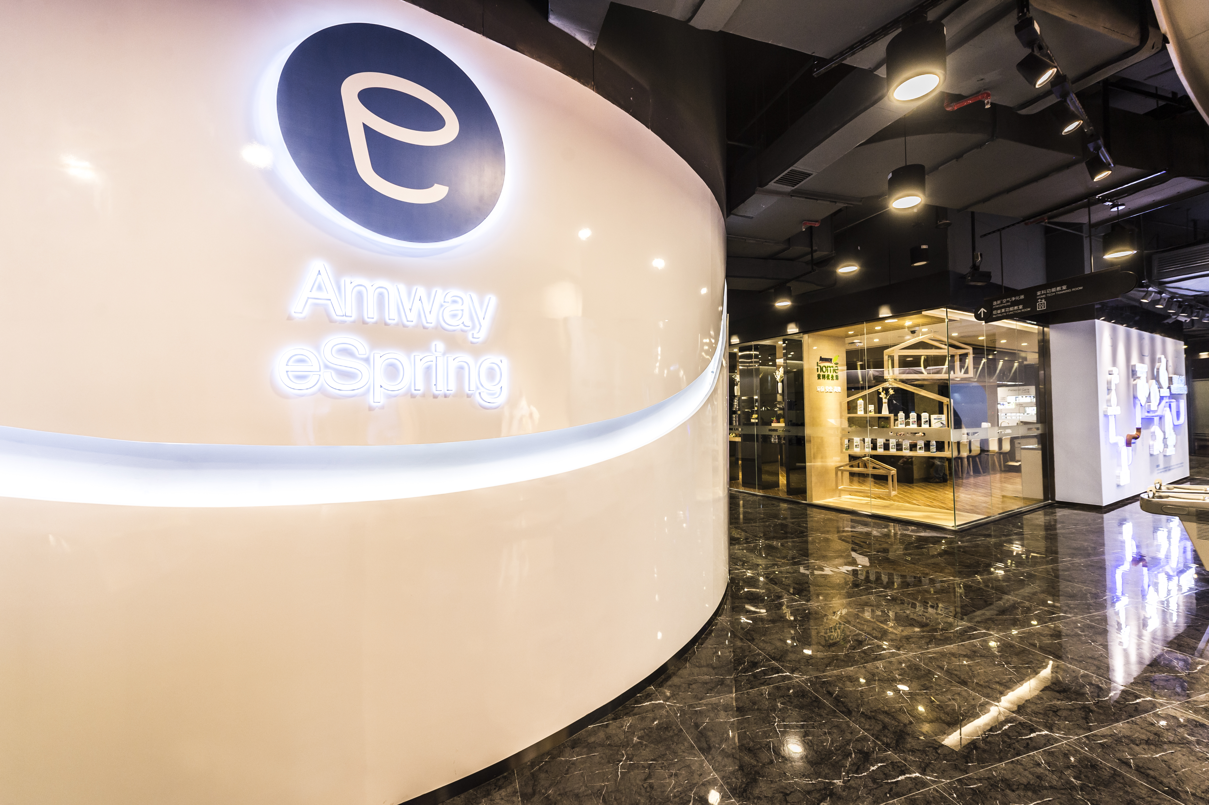 画面左侧是带有发光“Amway eSpring”标识的弧形展示墙，质感精致高级，右侧为玻璃围挡的安利家居科技体验展区，亮面大理石地面整洁光亮，整体空间现代大气，充分彰显安利高端专业的品牌调性，传递出科技感满满的家居健康服务理念。