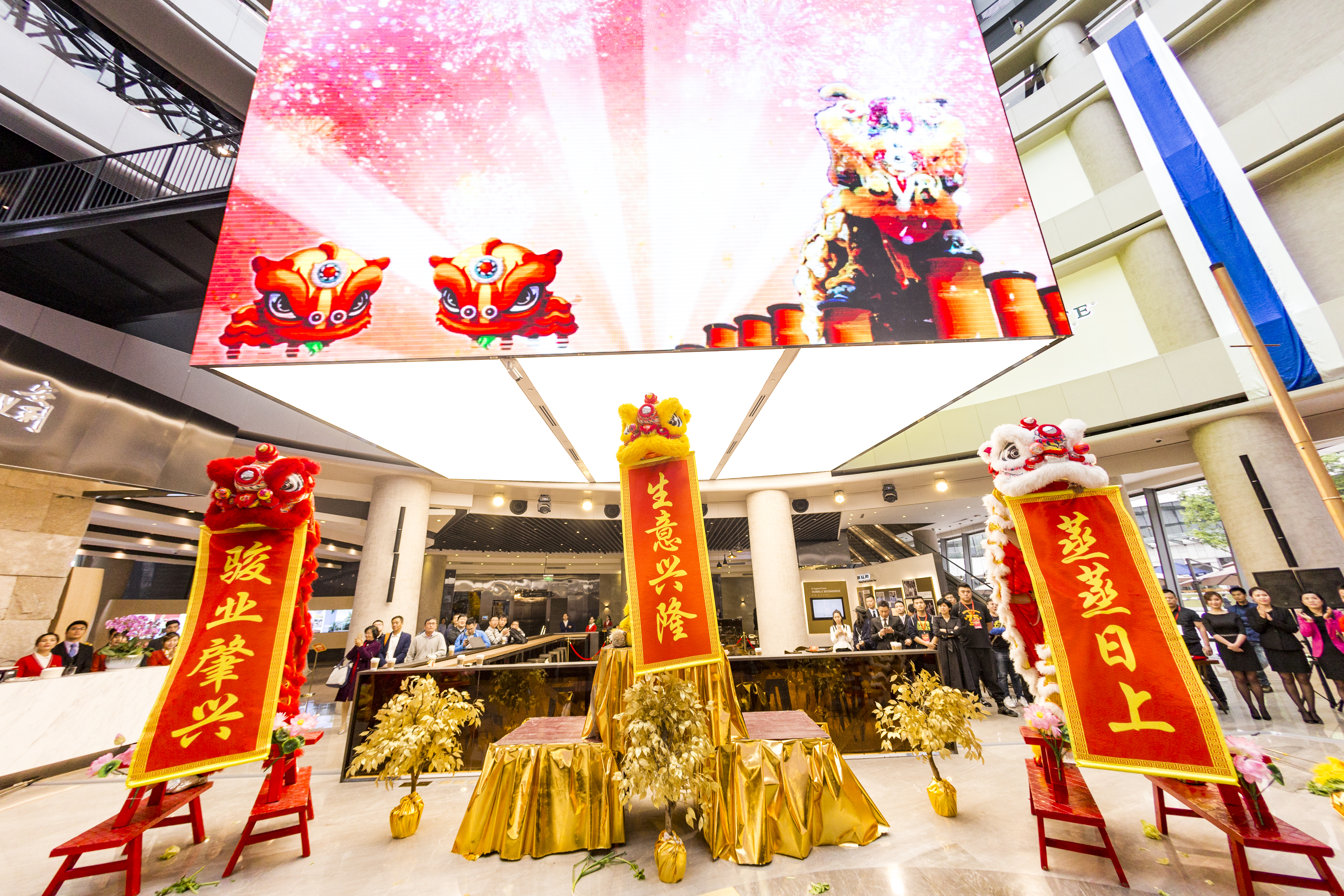 本次安利品牌庆典活动现场，三只醒狮分别持写有“骏业肇兴”“生意兴隆”“蒸蒸日上”的贺幅亮相，现场陈设金色装饰，上方大屏同步展示醒狮庆贺画面，众多参会伙伴驻足观礼，整体氛围喜庆热烈，尽显品牌向好发展的蓬勃朝气与美好期许。