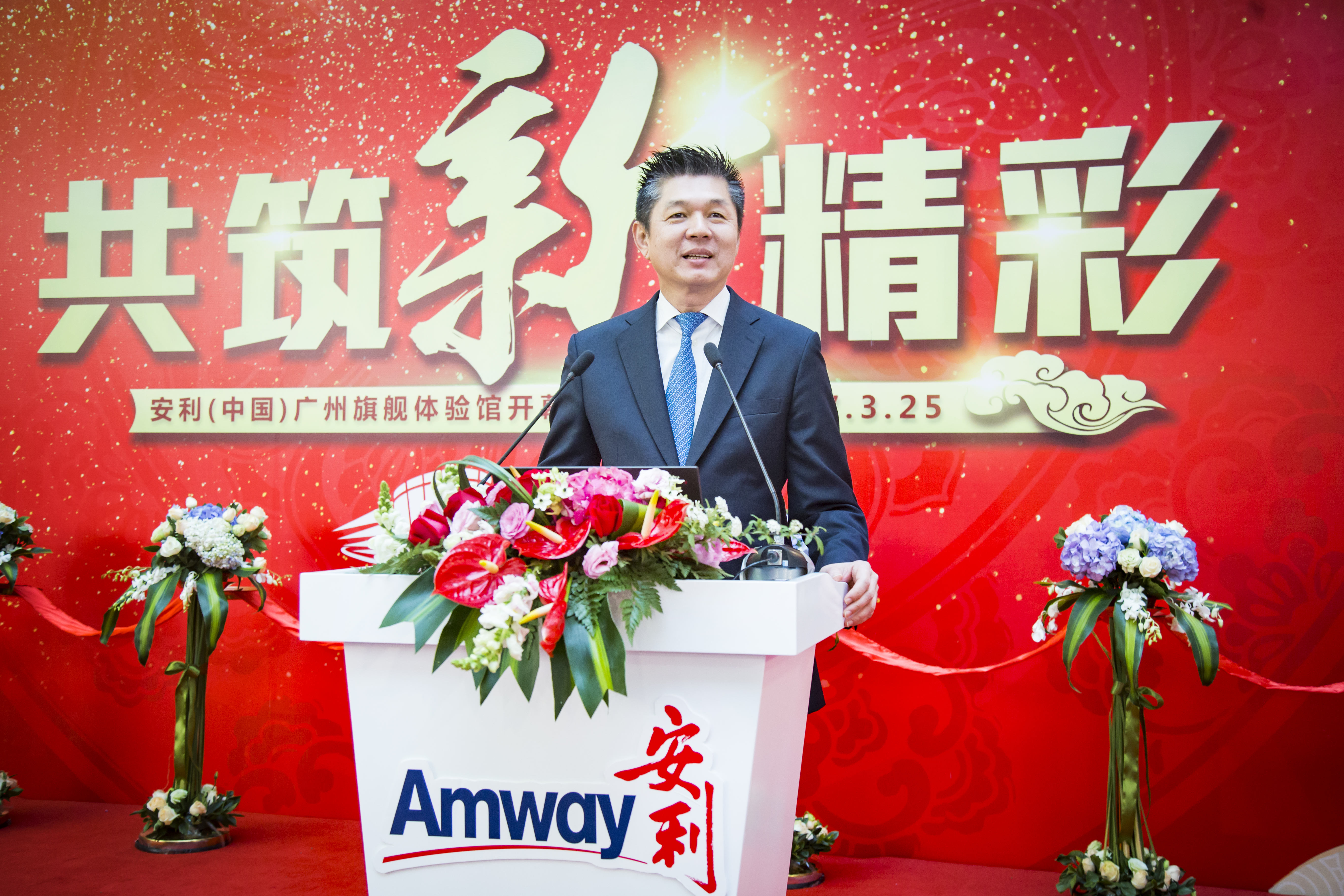 本次为安利（中国）广州旗舰体验馆开幕活动，红色背景板印有“共筑新精彩”主题字样，标注活动日期为7.3.25，氛围喜庆热烈。身着正装的男士站在装饰有鲜花、印有安利标识的致辞台后发言，现场庄重正式，尽显品牌积极向上的发展姿态。
