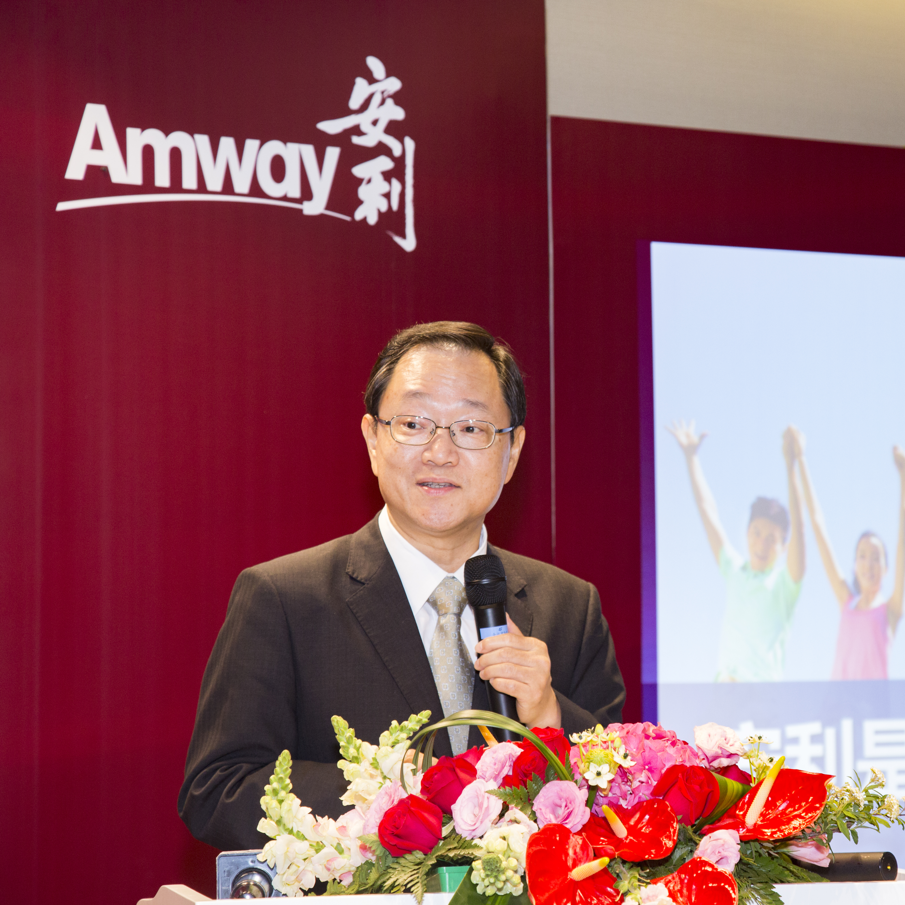 活动背景板印有核心标识“Amway安利”，一位身着正装、佩戴眼镜的男士手持话筒，在装饰有鲜切花的讲台发言，旁侧屏幕展示着充满活力的人物形象，整体氛围庄重专业，尽显安利活动积极正向的品牌风貌。