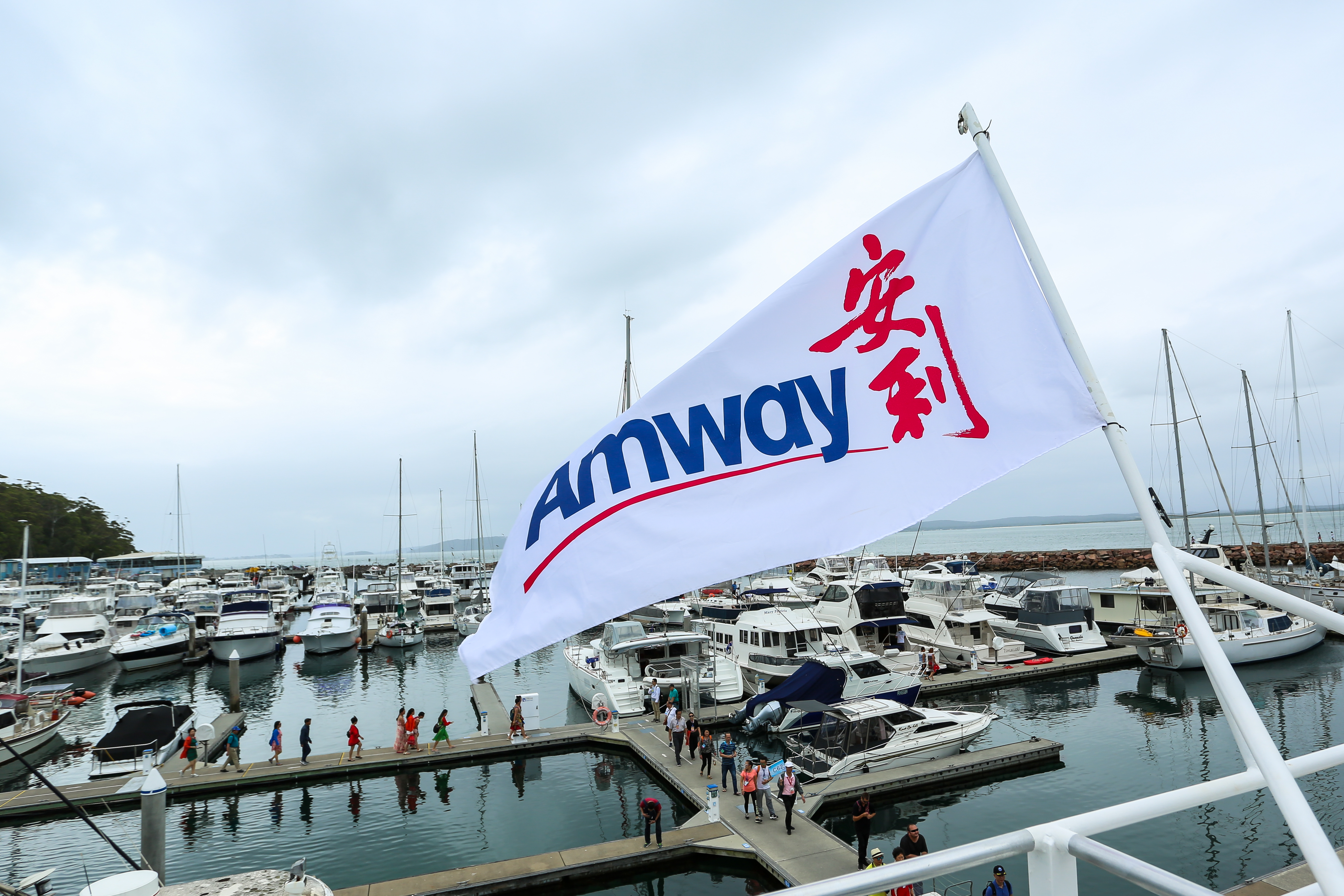 画面中印有“Amway”“安利”字样的品牌旗帜迎风飘扬，背景是停泊着众多游艇的开阔港口，水面澄澈，众多安利伙伴有序行走在泊位通道上，整体氛围轻松活力，尽显本次安利高端体验活动的惬意尊享感。