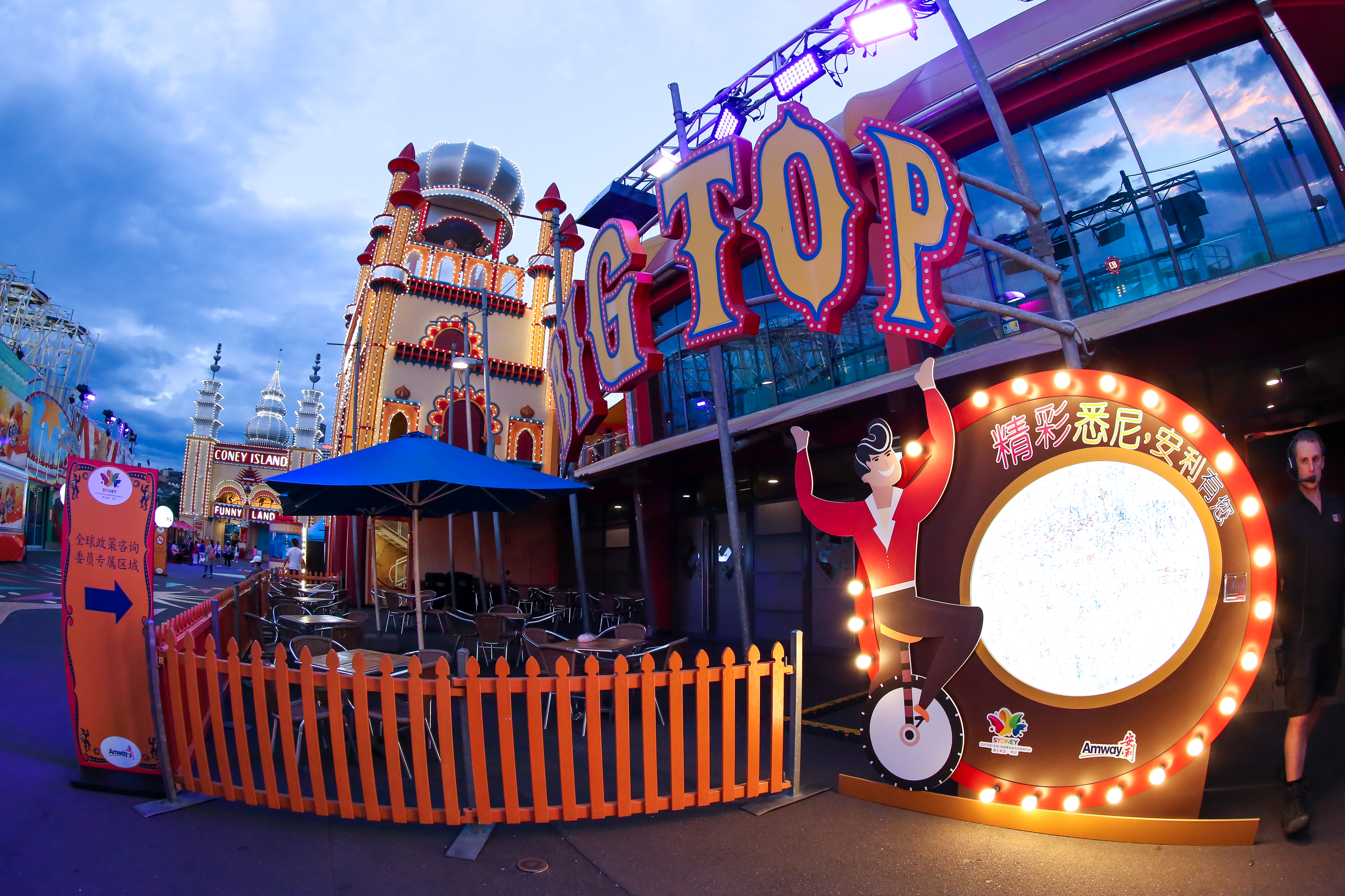 暮色下的游乐园专属活动区内，可见“BIG TOP”“CONEY ISLAND”园区标识，设有“全球政策咨询委员专属区域”指引牌，还有带有“精彩悉尼，安利有您”字样及Amway标识的打卡装置，场地配套休闲桌椅，布置明快活泼，氛围轻松愉悦，是安利为参会伙伴打造的专属活动空间。