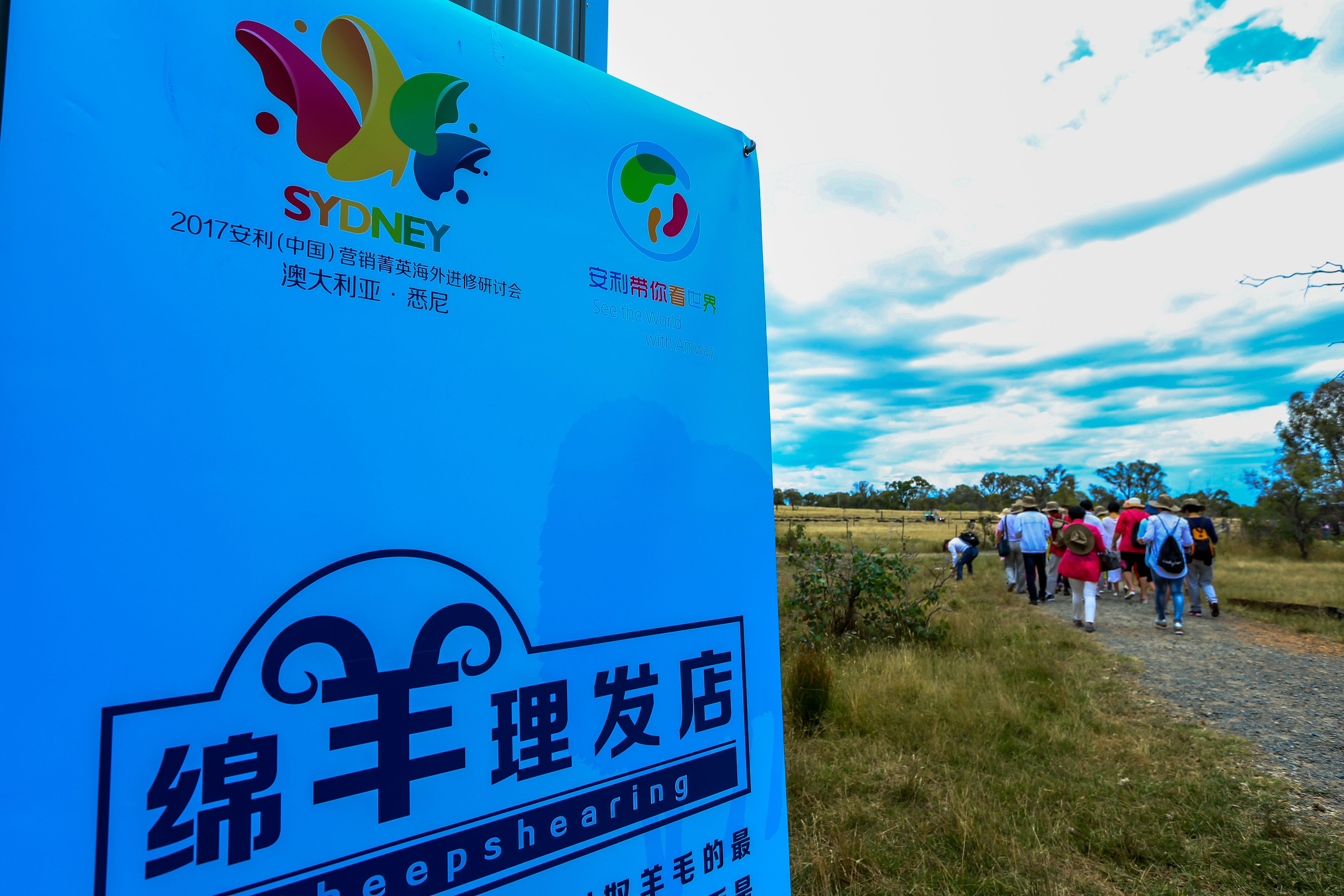 本次为2017安利（中国）营销菁英悉尼海外进修研讨会活动，现场设有标注“绵羊理发店”“安利带你看世界”的主题指示牌，参与活动的营销菁英正结伴沿郊外步道前行，旷野开阔天朗气清，氛围轻松鲜活，尽显本次海外进修的丰富趣味。