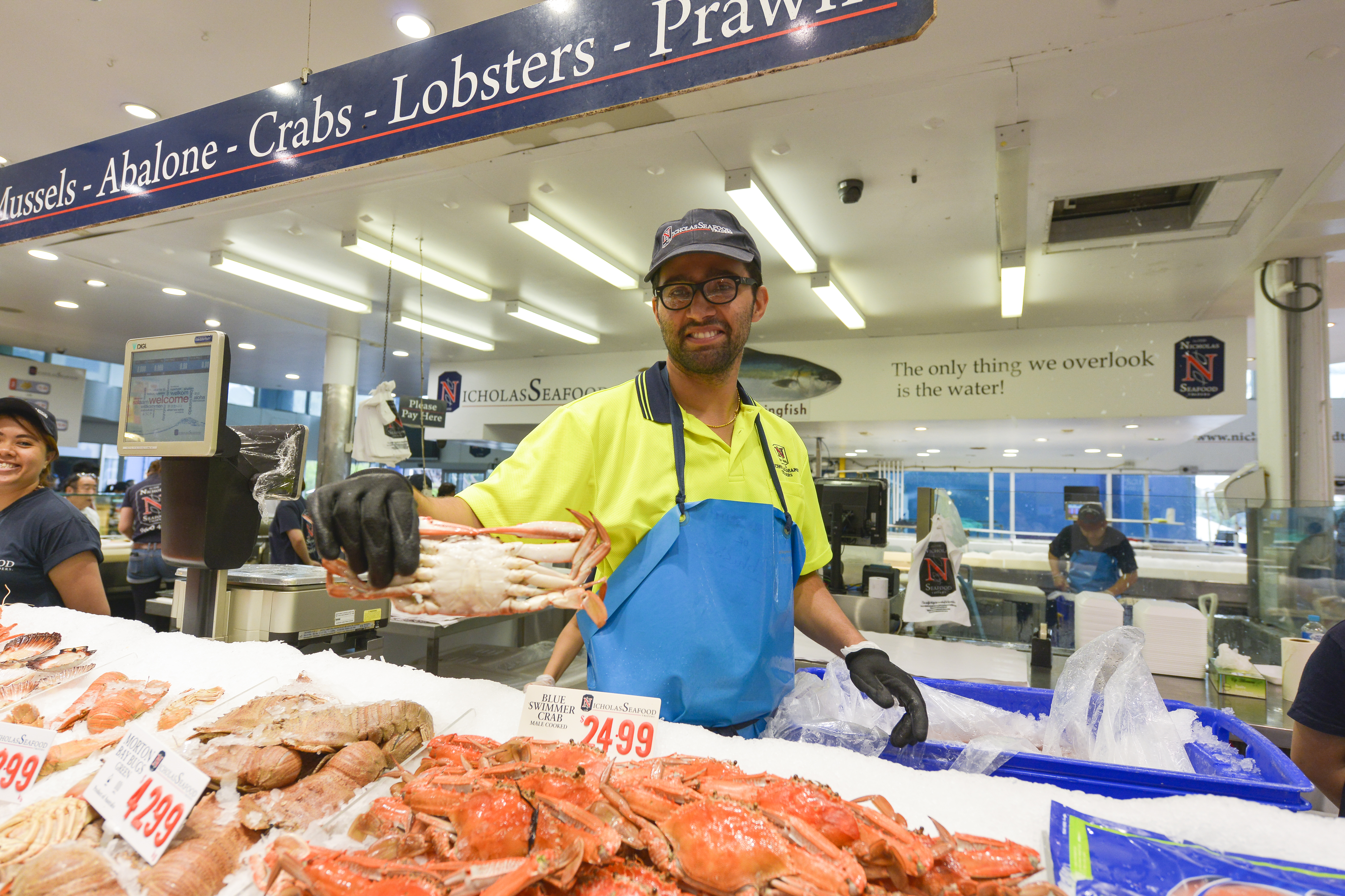 安利海外溯源团队探访Nicholas Seafood海鲜摊位，摊主面带笑容手持新鲜蓝泳蟹，冰台上摆放着品类丰富的鲜活海产，明码标注对应售价，摊位标语为“The only thing we overlook is the water!”，现场氛围鲜活热闹，尽显海产品质优势。