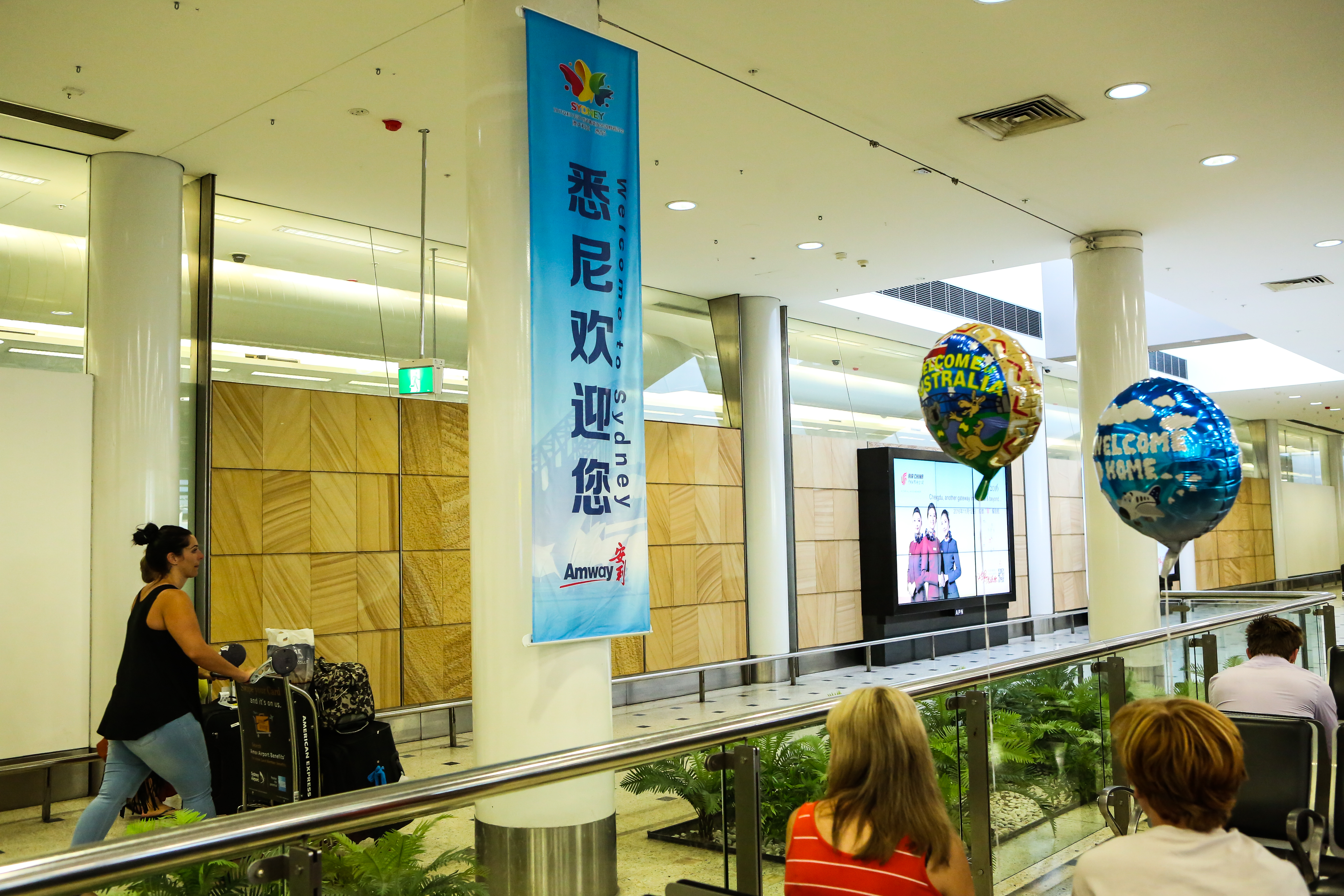 画面为悉尼机场内部场景，立柱悬挂印有“悉尼欢迎您”“Welcome to Sydney”及“Amway安利”标识的蓝色道旗，场内搭配印有“WELCOME AUSTRALIA”“WELCOME HOME”字样的主题气球，环境明亮整洁，氛围热烈暖心，彰显对安利活动参会人员的欢迎诚意。