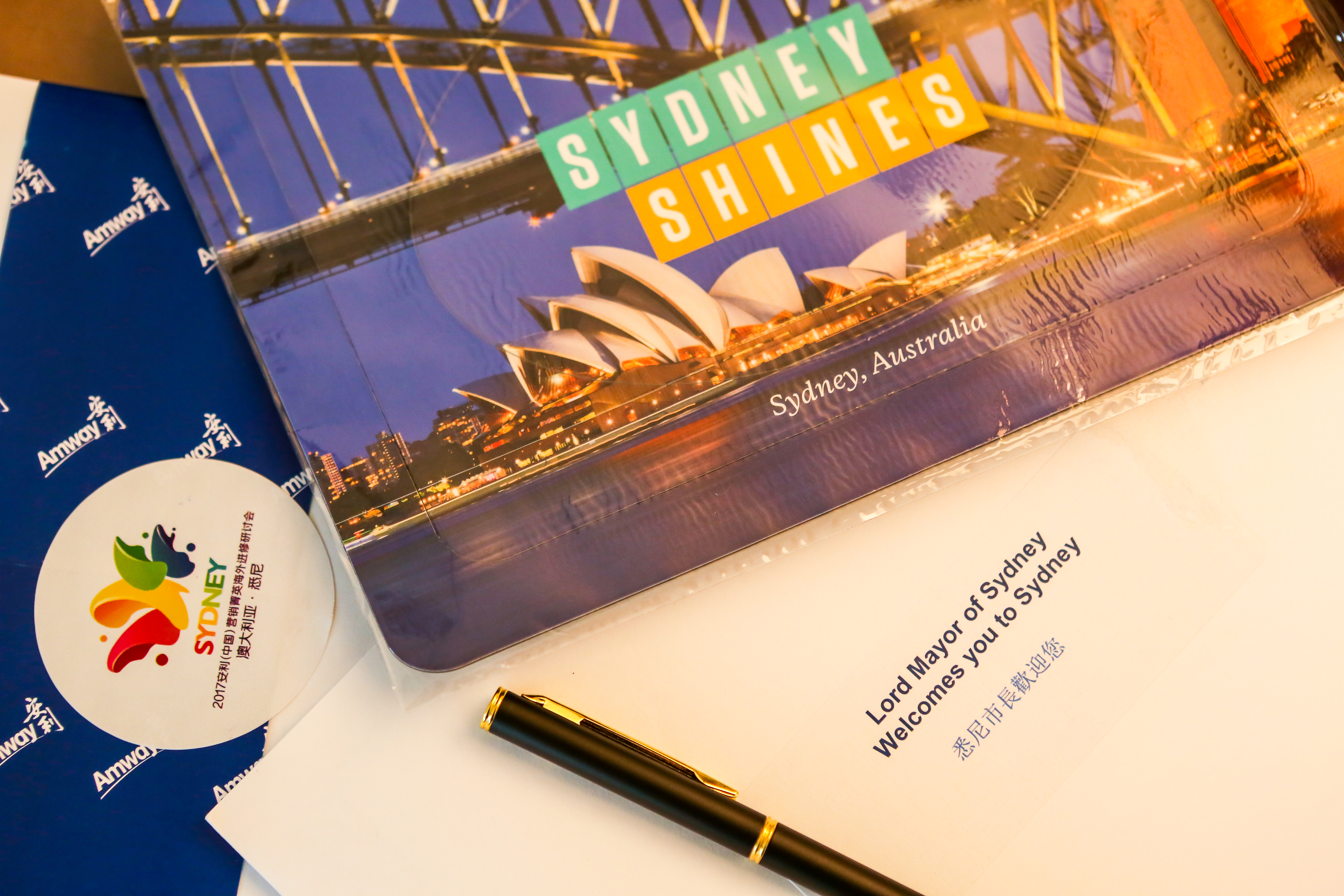 画面展示本次活动相关配套物料，印有悉尼歌剧院、海港大桥夜景与“SYDNEY SHINES”字样，包含活动专属标识、悉尼市长欢迎信函及安利品牌周边，质感精致考究，尽显本次海外进修活动的专属礼遇与隆重氛围。