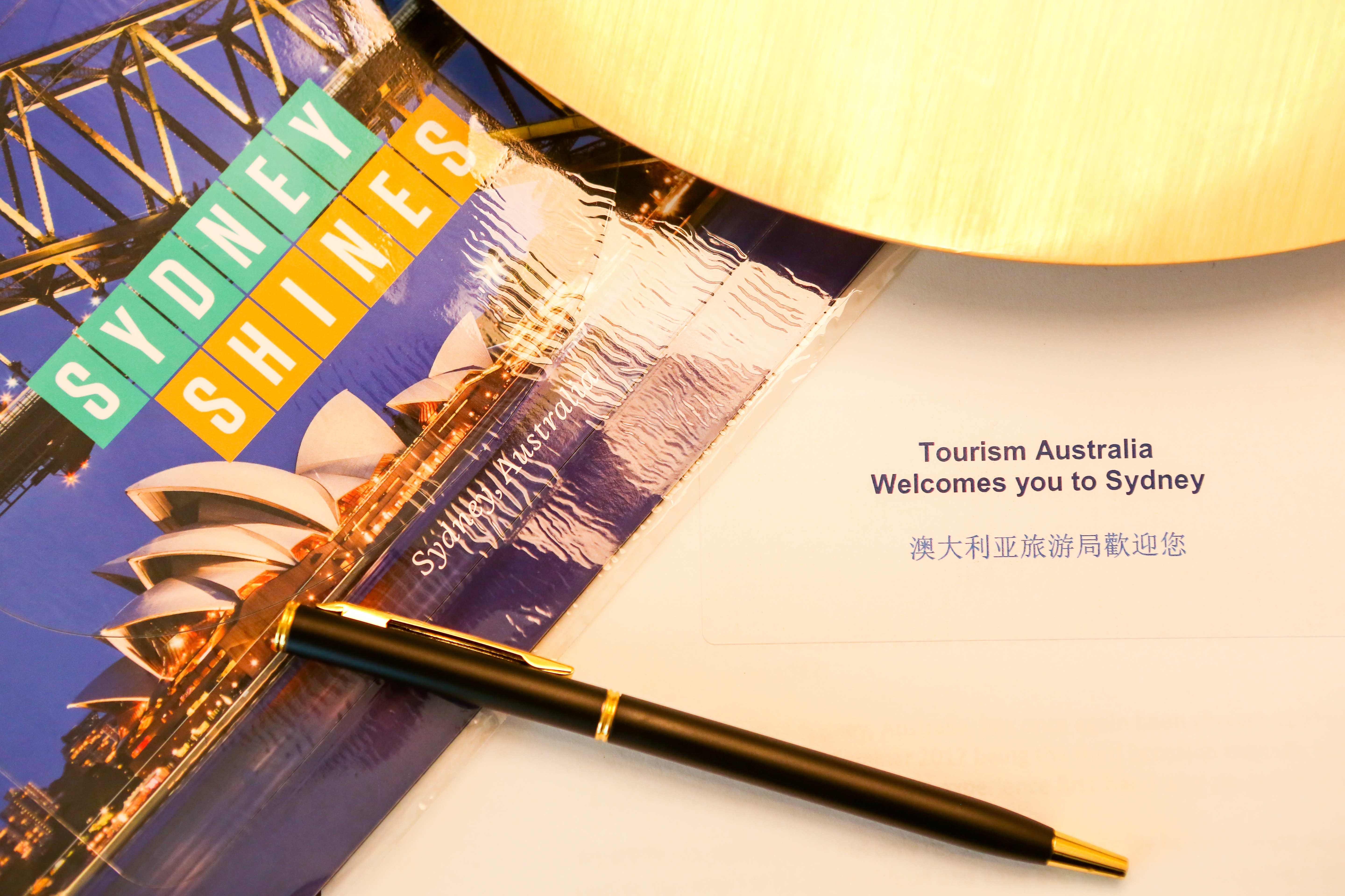 画面呈现印有悉尼歌剧院、海港大桥，标注“SYDNEY SHINES”的主题宣传册，搭配印有“Tourism Australia Welcomes you to Sydney”“澳大利亚旅游局欢迎您”的官方欢迎函，旁置黑金质感签字笔，整体精致正式，尽显本次悉尼活动的专属礼遇感。