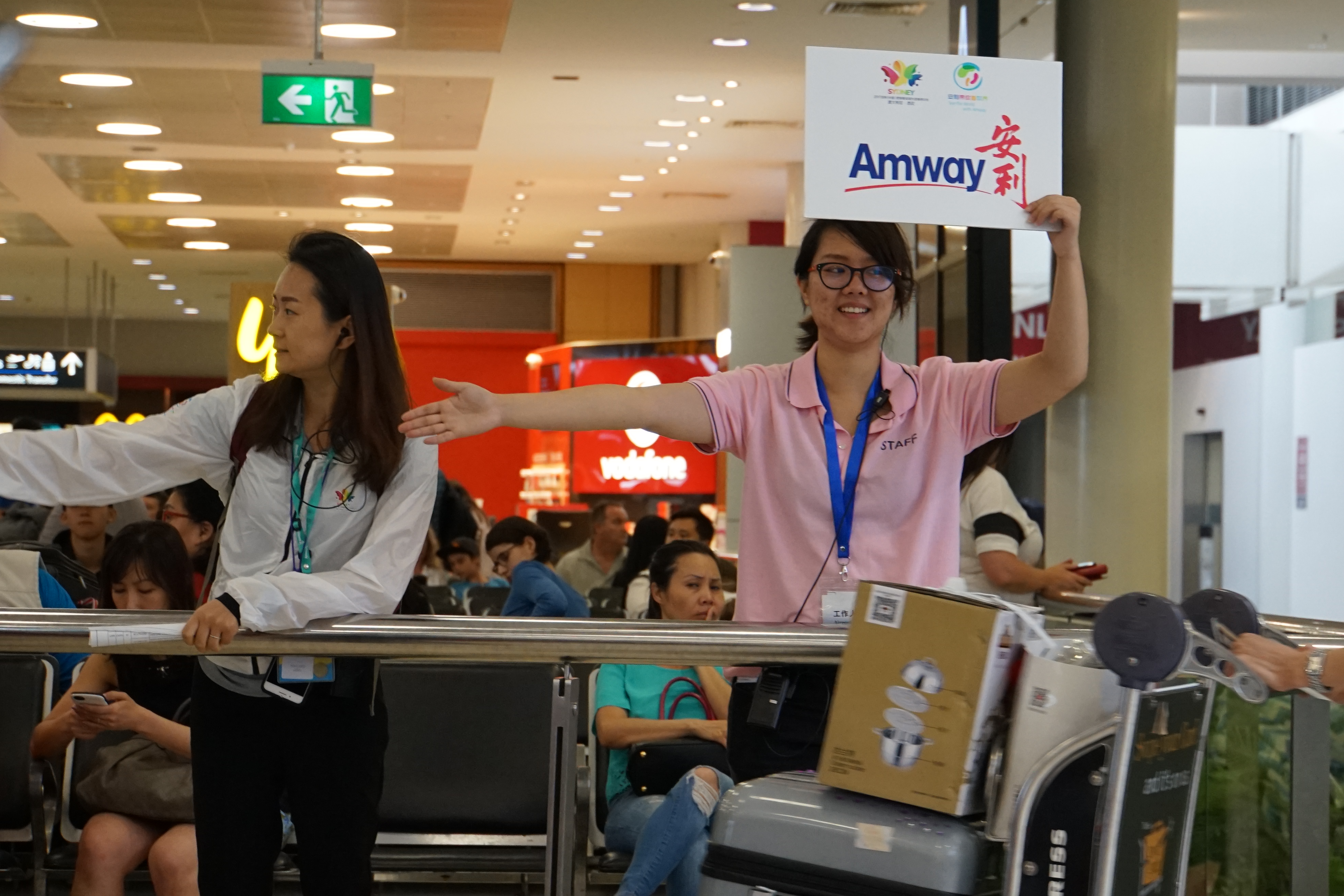 机场接待现场，安利工作人员身着工服，面带笑容高举印有“Amway安利”字样的指引牌，伸手引导前来的参会伙伴，接待流程有序推进，氛围热情周到，充分展现安利细致贴心的活动服务风貌。