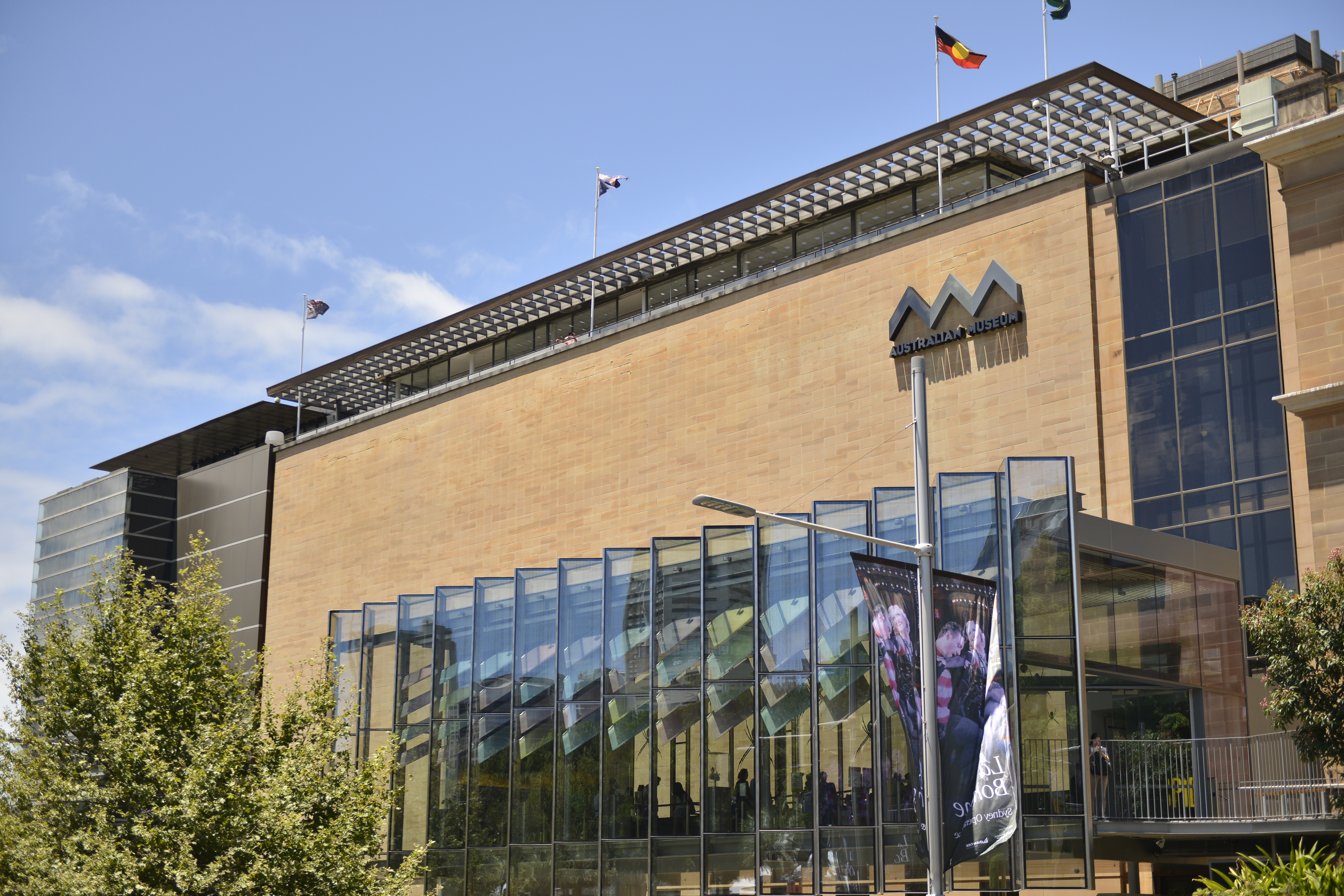 画面为晴朗天气下的澳大利亚博物馆（AUSTRALIAN MUSEUM），暖调砂岩外墙搭配通透玻璃入口，建筑顶部悬挂多面旗帜，周边绿植鲜亮，整体开阔明朗，是本次安利澳洲活动的举办场地。