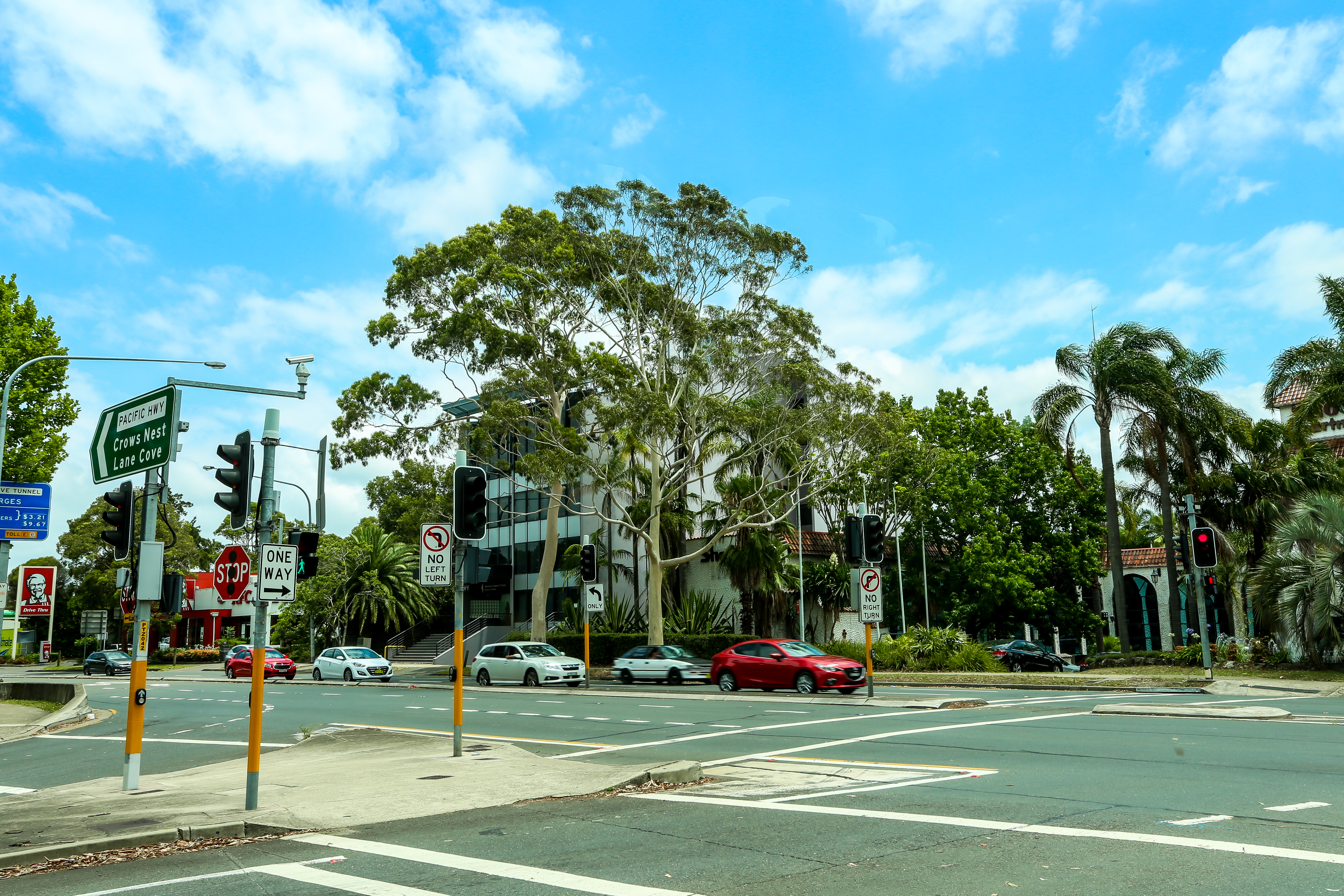 画面为晴朗天气下的城市路口，可见“PACIFIC HWY”路牌、“STOP”“ONE WAY”等交通标识，往来车辆有序通行，路旁绿植葱郁、建筑整洁，氛围闲适明快，尽显安利海外研讨会举办地舒适宜人的城市风貌。