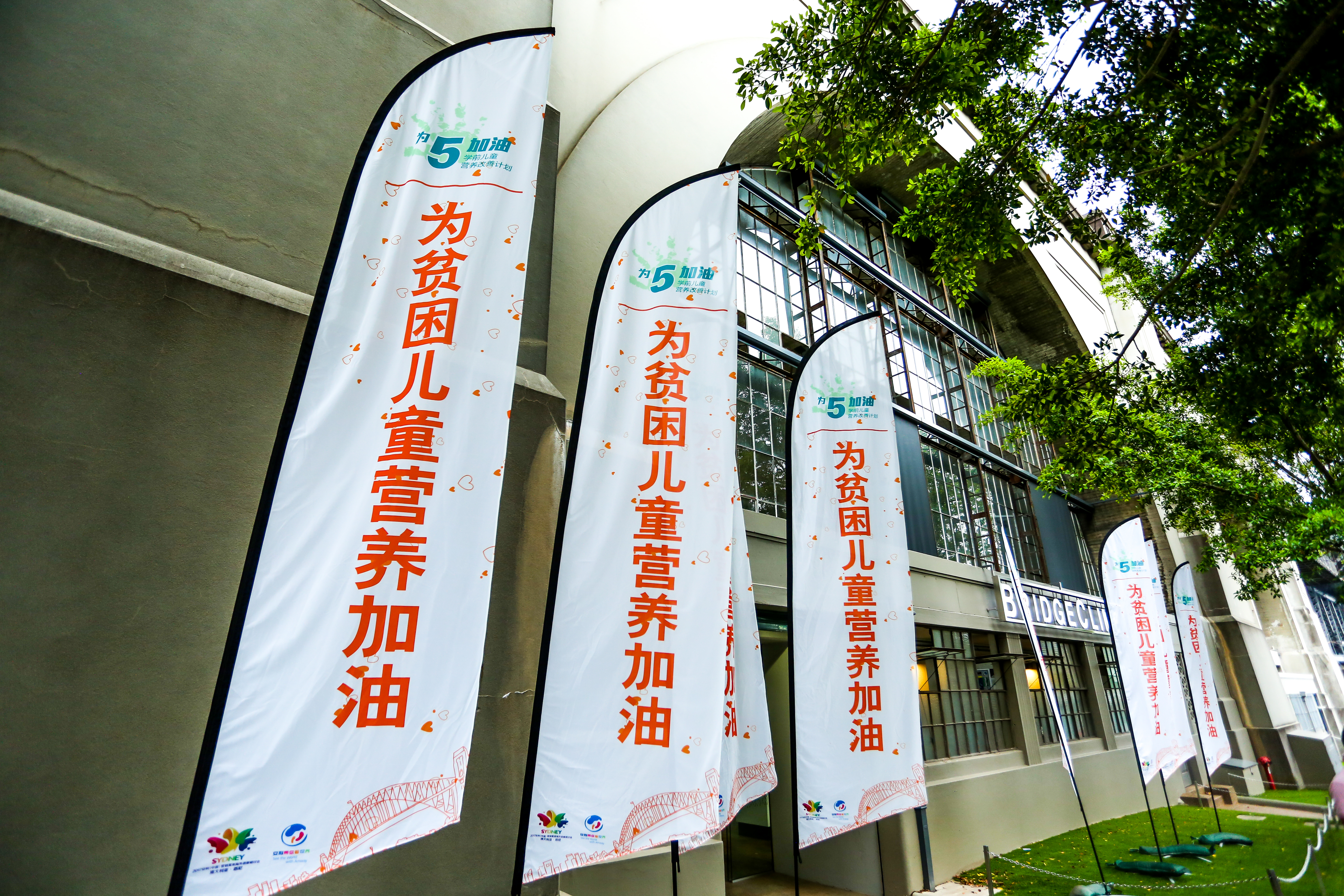 画面呈现安利“为5加油”学前儿童营养改善计划活动现场，多面印有“为贫困儿童营养加油”字样的道旗沿建筑外整齐排布，周边绿植葱郁，氛围温暖正向，充分彰显安利投身儿童公益、助力贫困儿童营养改善的责任初心。