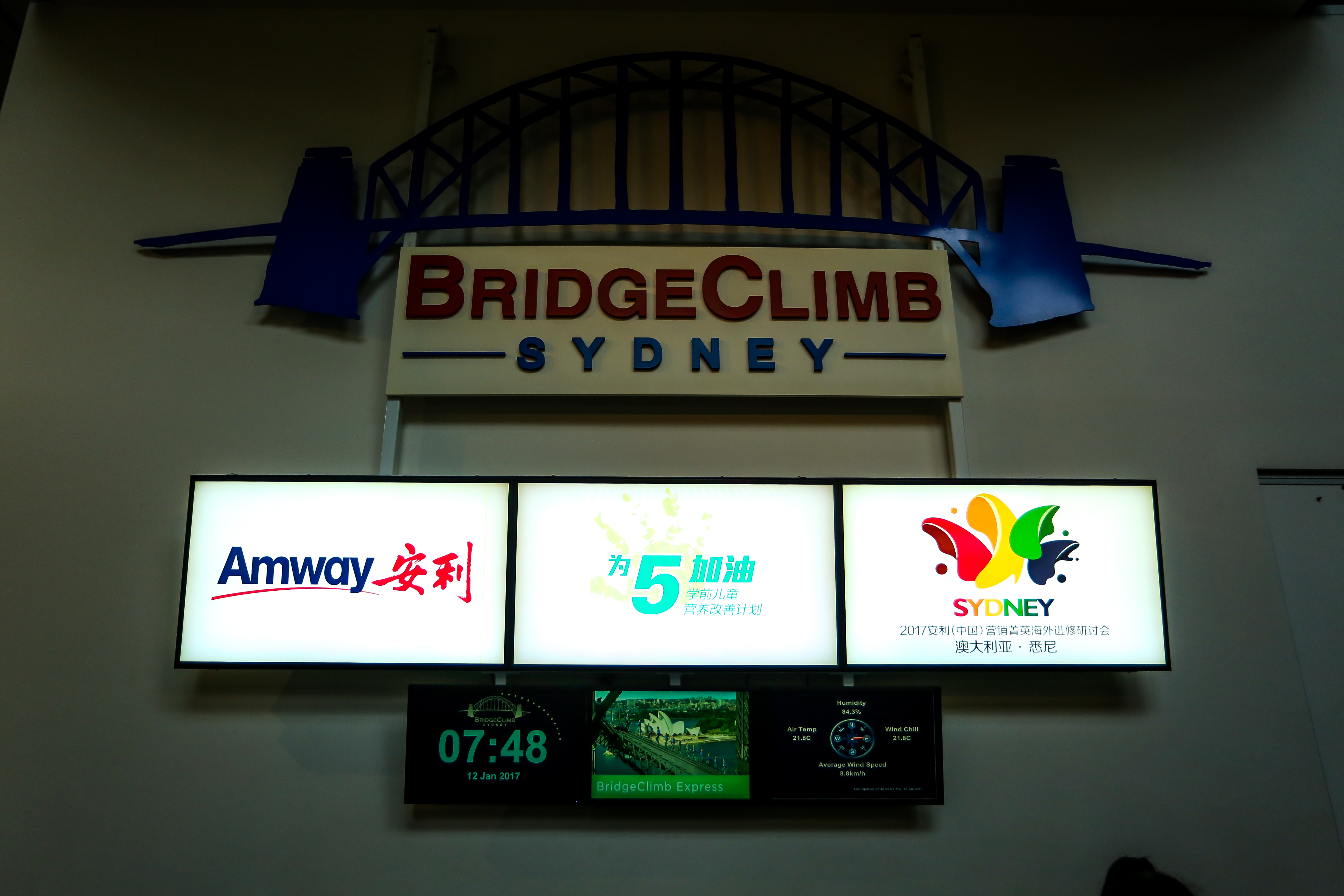 画面顶部是悉尼海港大桥造型的“BRIDGECLIMB SYDNEY”标识，下方灯箱依次展示Amway安利品牌标识、“为5加油 学前儿童营养改善计划”标识、2017安利（中国）营销菁英海外进修研讨会悉尼站标识，底部配套显示时间、天气信息，整体清晰传递活动专属属性，正式明快。