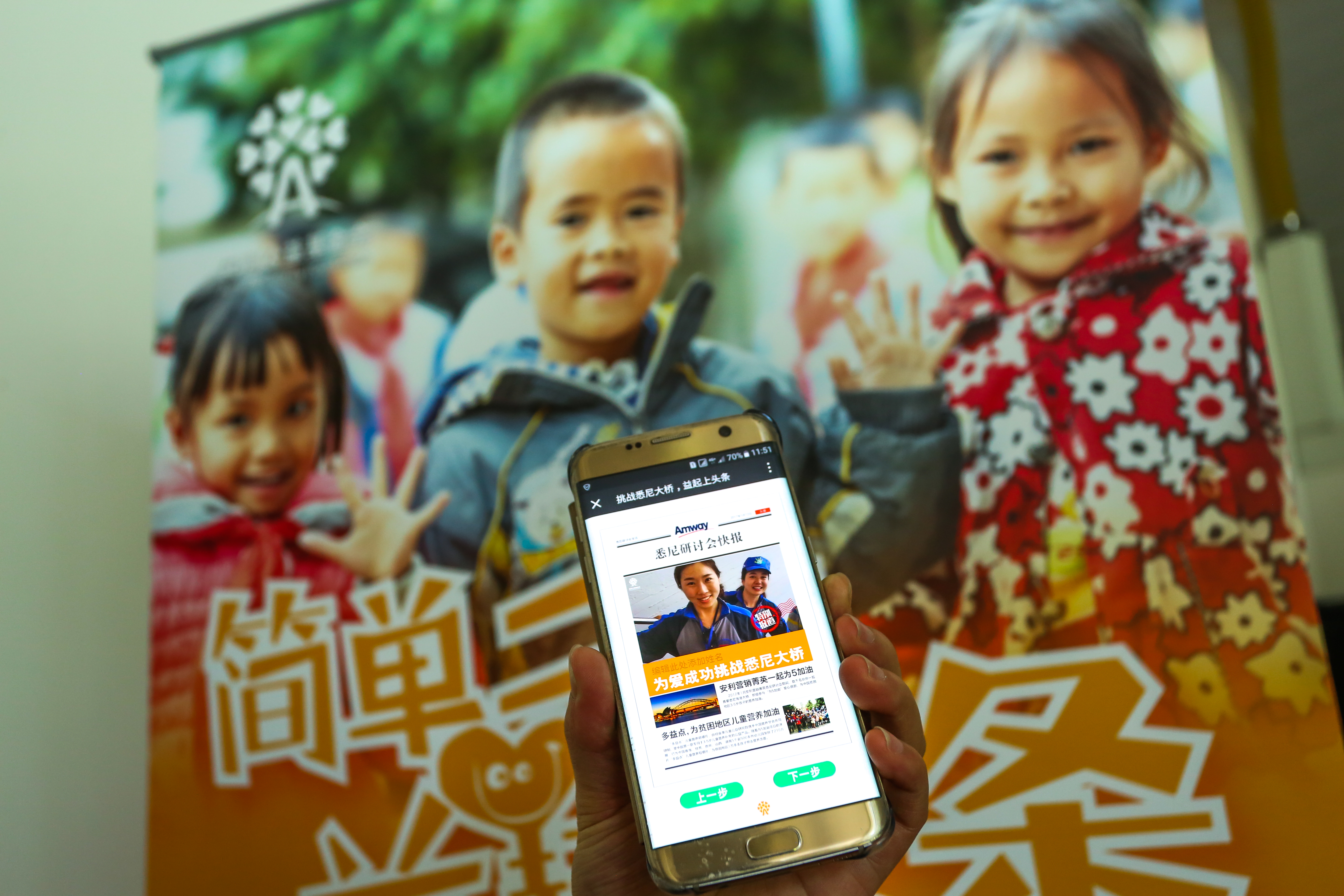 画面中有人手持手机展示安利悉尼研讨会快报页面，页面包含“为爱成功挑战悉尼大桥”“安利营销精英一起为5加油，为贫困地区儿童营养加油”等内容，背景是印有孩童笑脸的安利公益海报，整体氛围温暖正向，彰显安利践行公益的品牌温度。