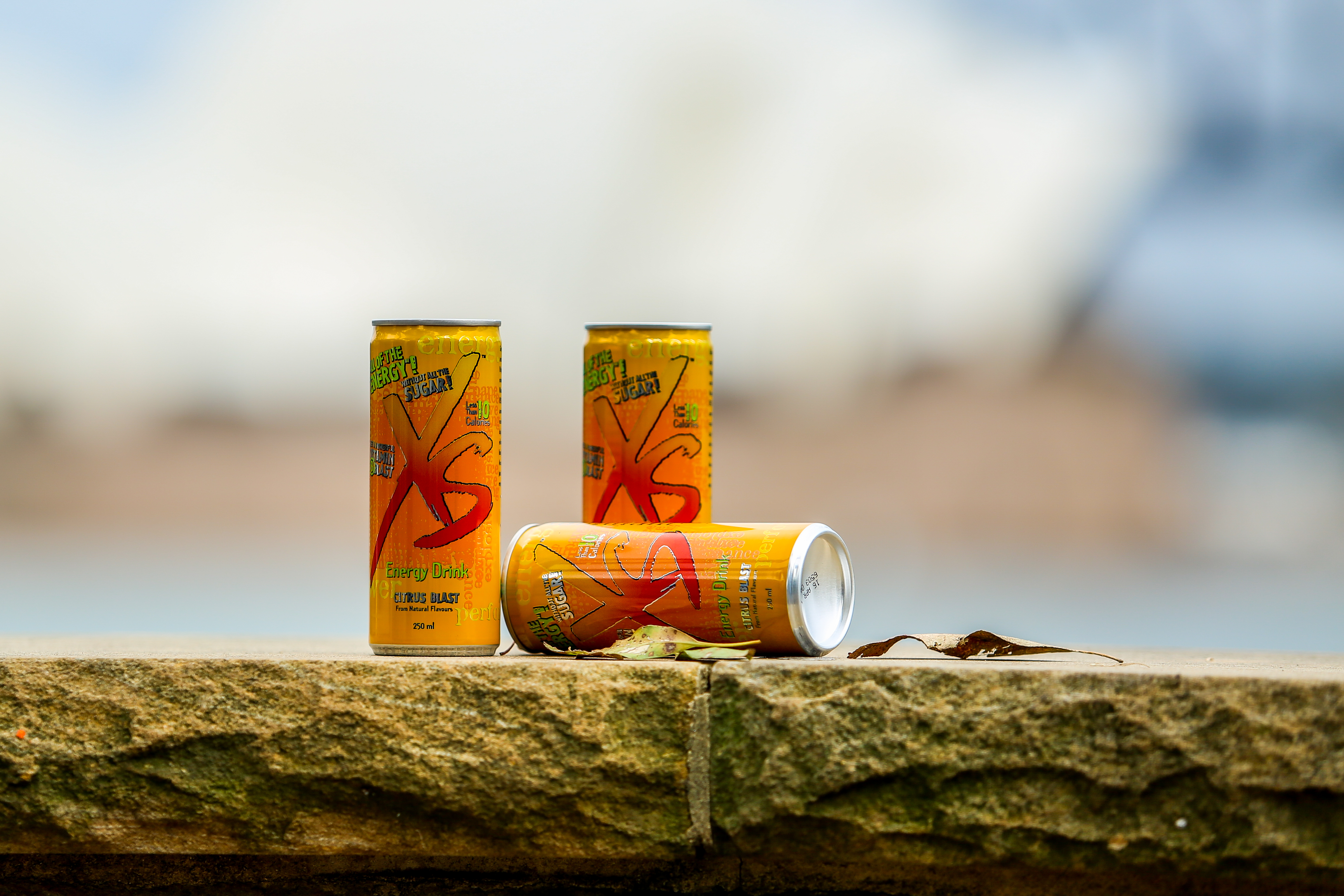 画面呈现三罐安利XS柑橘味能量饮料，罐身印有“XS”“Energy Drink”“CITRUS BLAST”“NO ADDED SUGAR”等标识，产品摆放于户外石质台面上，背景是虚化的开阔自然景观，氛围清爽鲜活，传递出产品健康活力的属性。